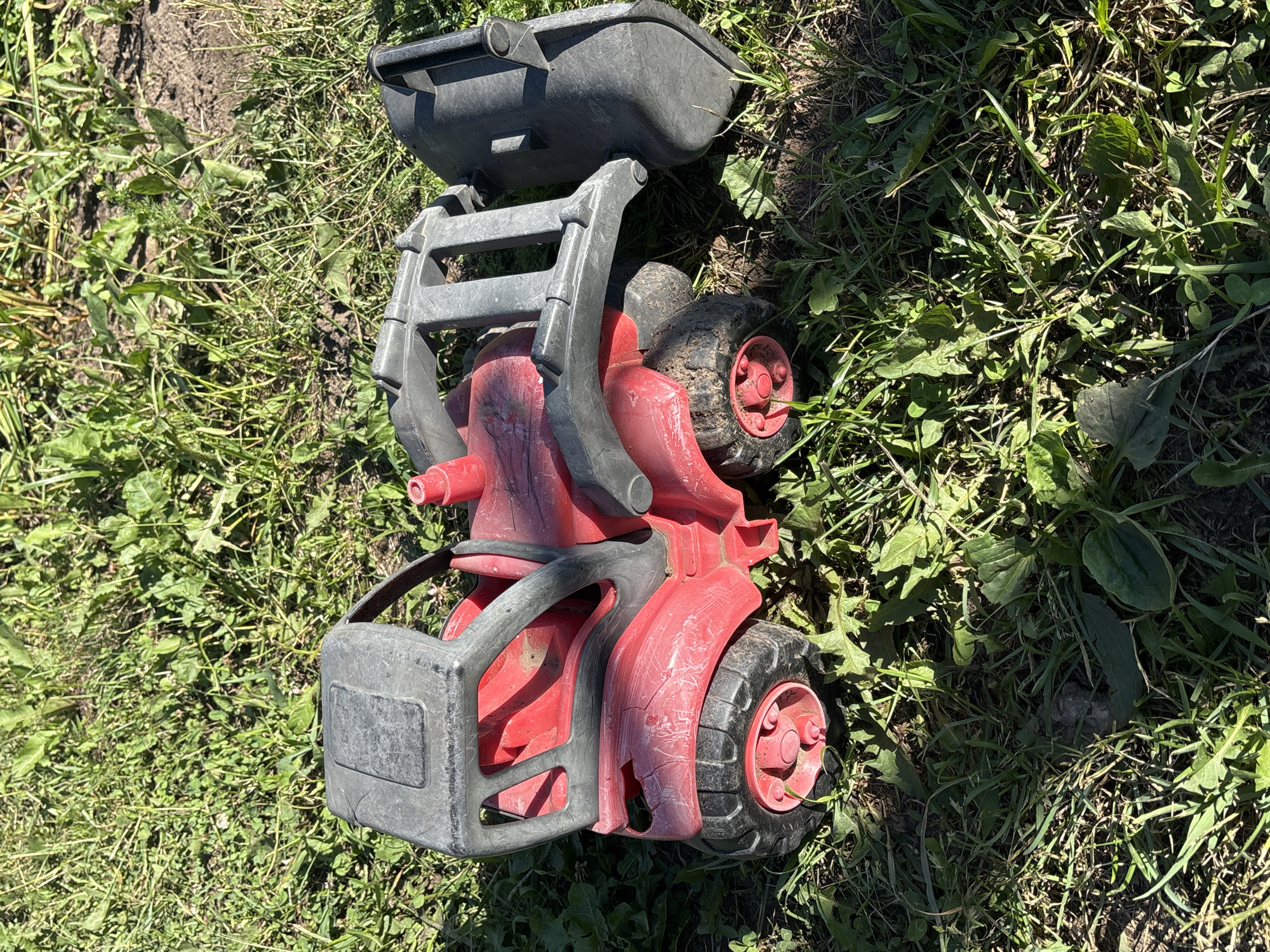 A red toy tractor with a gray loader is lying on its side in a grassy area. The tractor is plastic and appears to be old and somewhat damaged, with some cracks visible on the body. The loader is raised and angled forward. The ground is covered in green grass and some dry dirt. The image is taken from a slightly elevated angle, looking down on the toy. There are no people or animals visible in the image. The lighting suggests it is daytime, likely outdoors. The provided location of Новый Кинер, Russia, does not offer specific environmental context visible in the image itself.