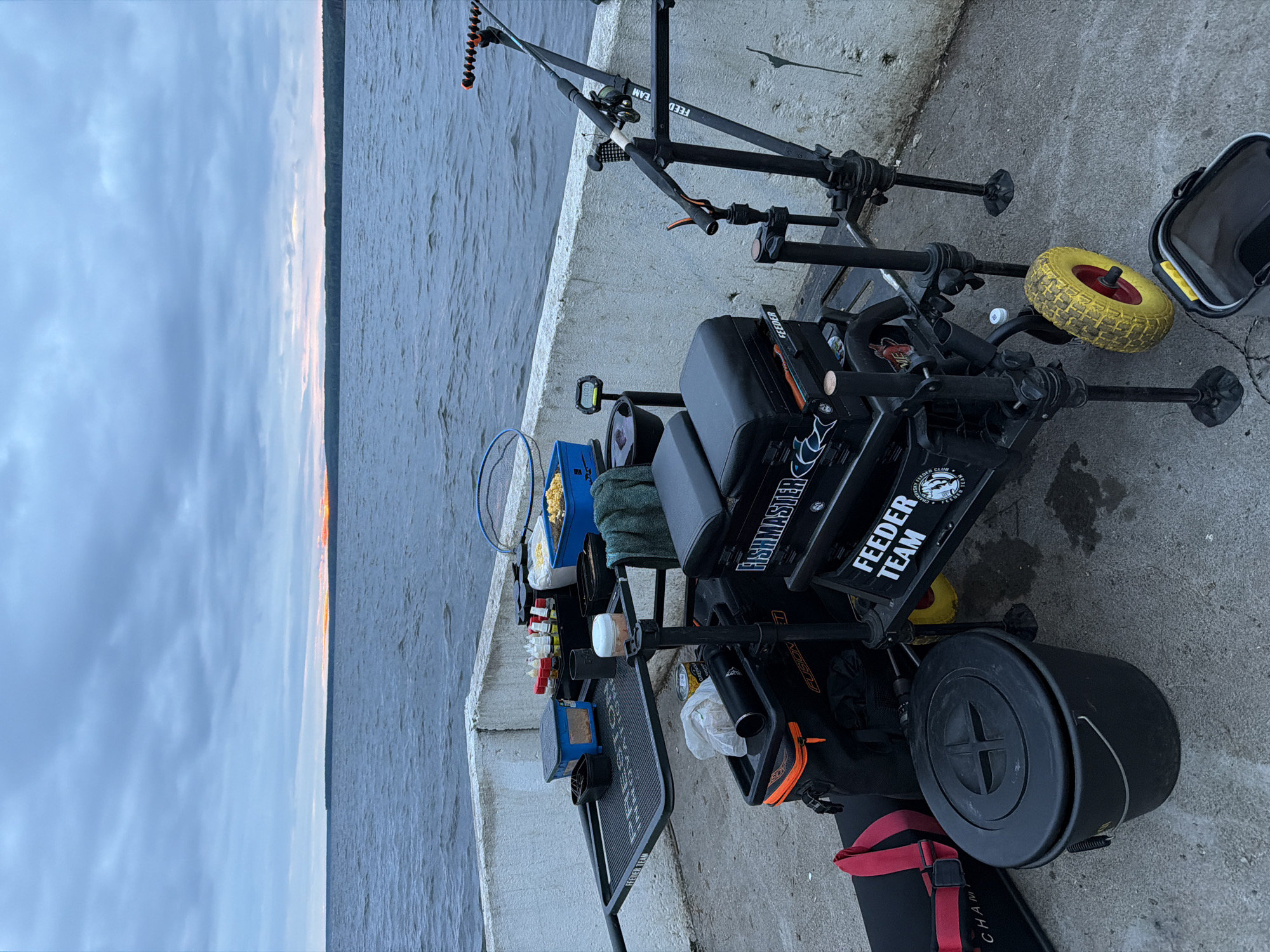 The image displays a comprehensive fishing setup situated on a grey concrete embankment adjacent to a large body of water in the city of Chaikovsky, Russia. The sky is uniformly overcast with layers of grey clouds, and a narrow band of orange and yellow light on the distant horizon indicates either sunrise or sunset, positioned above a dark treeline. The water surface exhibits ripples and small waves, suggesting movement.
In the foreground, a professional-grade black fishing seat box, labeled "FISHMASTER" and "FEEDER TEAM," is centrally positioned. It features a padded black seat cushion, multiple adjustable black legs with wide mud feet, and a yellow wheel with a red hub attached to its right side for transport. A black side tray with a textured surface, branded "PRESTON FEEDER TEAM," is affixed to the left of the seat box. On this tray, a small blue rectangular container, a black cup, a white container with a lid, and an organizer holding multiple small transparent bottles are neatly arranged. A dark green towel hangs nearby. Below the tray, a black soft bag with an orange zipper, marked "FUSION," is visible. Additionally, a blue rectangular container filled with a yellow granular substance is placed near the seat box.
To the right of the seat box, a dark-colored fishing rod with an orange tip protector and a dark fishing reel is mounted on a multi-adjustable black rod rest system, which also displays "FEEDER TEAM" branding. A small digital scale, reading "0.00," is attached to one of the vertical supports of this system. A blue fishing net with a dark handle is partially visible, leaning against the concrete embankment wall behind the rod setup.
On the concrete ground to the left, a black rectangular bag with red straps, partially showing "CHAM," lies horizontally next to a black bucket with a lid featuring a cross-shaped design and a metal handle. Another black bucket with a yellow rim and handle is situated near the yellow wheel of the seat box. No individuals are present in the scene.