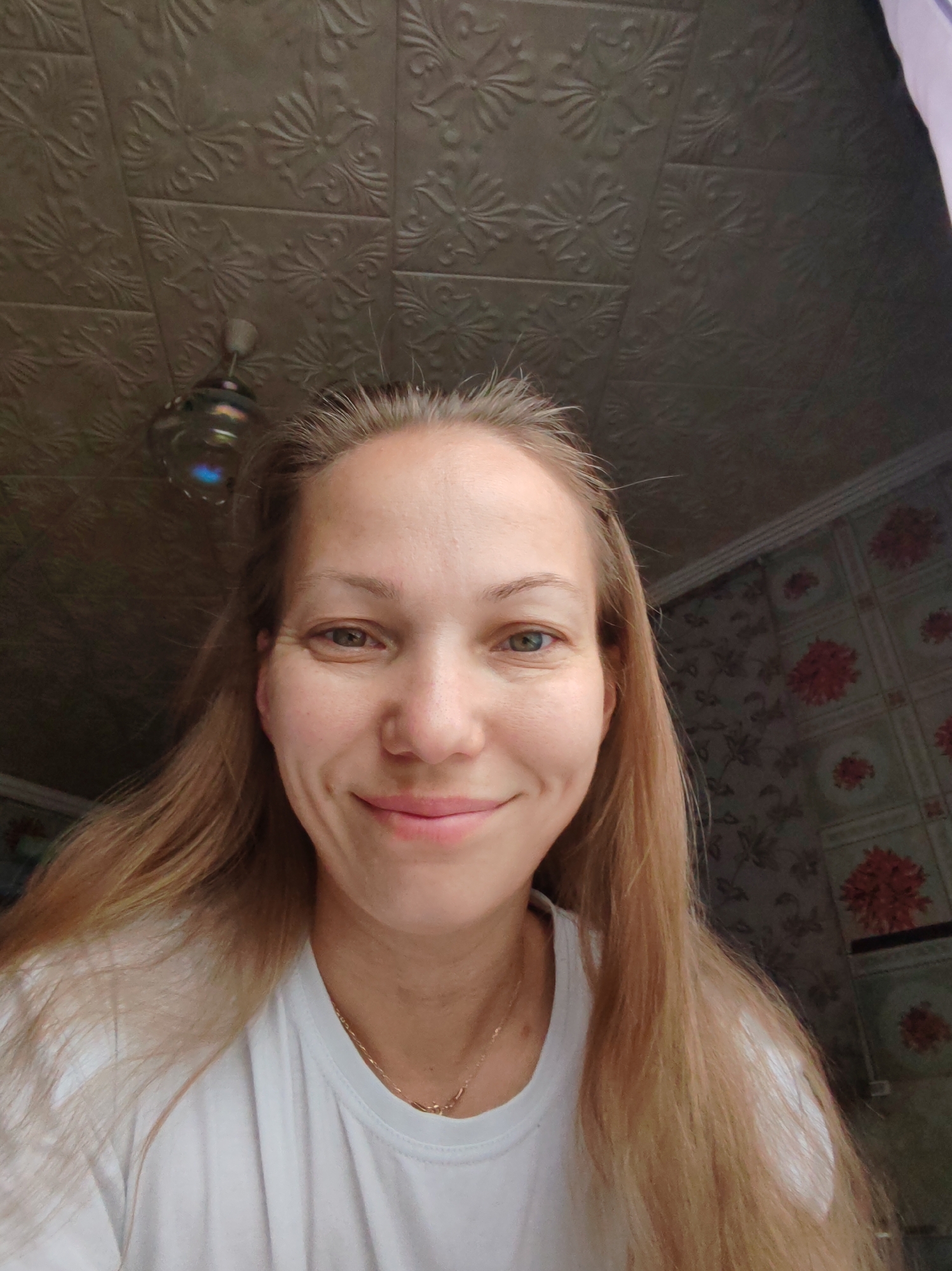 A blonde woman with light skin and blue eyes is looking directly at the camera with a slight smile. She has long, straight blonde hair that extends past her shoulders. She is wearing a white t-shirt with a delicate gold necklace. The background appears to be an indoor setting, possibly a room with textured walls and a patterned wallpaper featuring red floral designs. A ceiling fixture is visible above and to the left of the woman's head. The lighting is soft, casting a gentle glow on her face. The image is a close-up, focusing on the woman's upper body and face. There is no indication of the location being in Чухлэм, Russia.