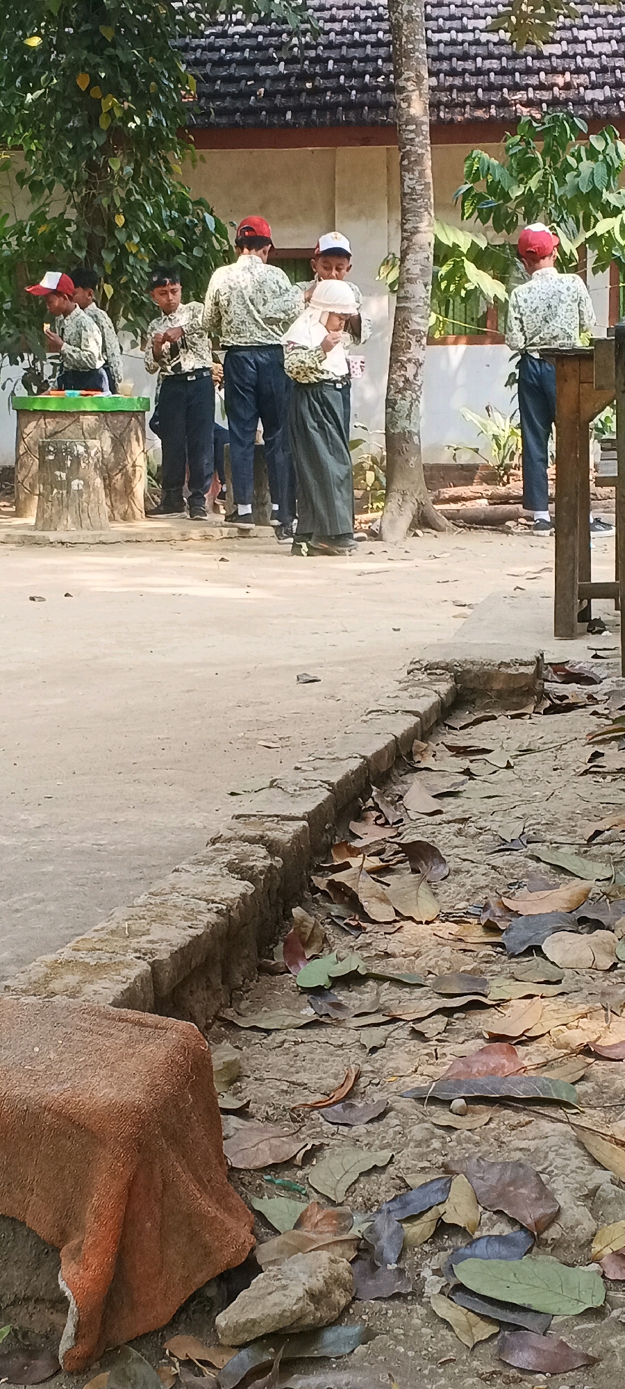 In Penanggungan, Indonesia, a group of five school children, three boys and two girls, are gathered outdoors near a building with a tiled roof. The children are wearing school uniforms, consisting of patterned shirts, dark pants, and caps. Two of the boys are wearing red caps, one boy has a white and red cap, and the girls appear to be wearing headscarves. They are standing on a dirt path, and there are fallen leaves scattered on the ground. In the foreground, a low, stone border runs along the edge of the path, with a roughly shaped orange object placed on it. A tree trunk is visible to the right of the children. The children appear to be interacting with each other, with some holding cups. The scene suggests a break or activity taking place during the school day in an outdoor setting.