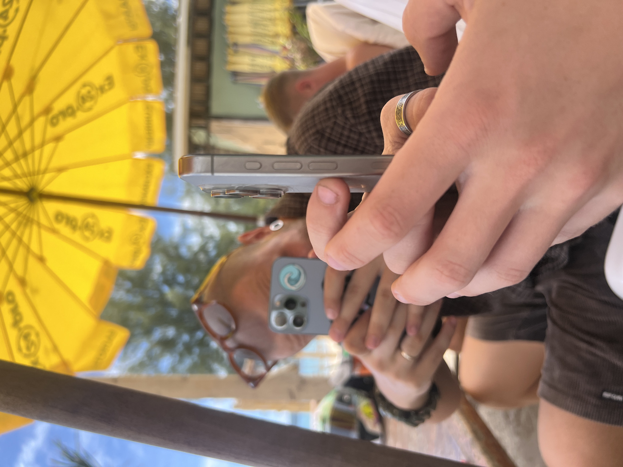 An individual, seen from the chest up, holds a smartphone in their right hand, with their left hand also in the frame. They are wearing glasses and have short hair. The phone is angled upwards, suggesting it is being used to take a photo. In the background, another person is visible, partially obscured by the foreground subject. The scene appears to be outdoors, with a bright yellow umbrella providing shade. The environment suggests a tropical location, possibly a beach or resort setting, with some foliage and structures visible in the distance. The lighting indicates it is daytime, with the sun shining through the umbrella. The overall impression is a candid moment captured on a mobile device, likely in a leisure setting. The location is Choeng Thale, Thailand.