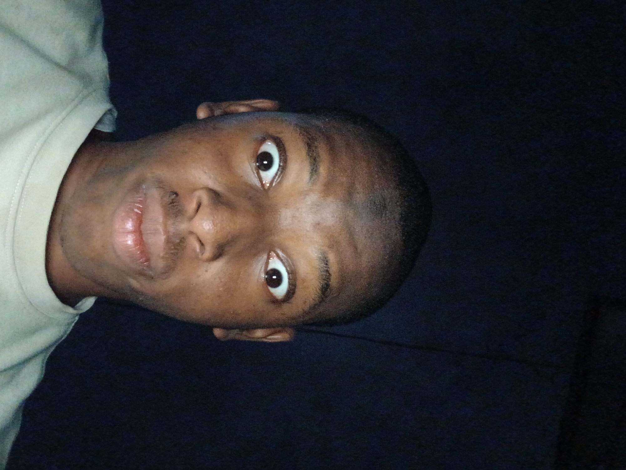 A dark-skinned man is photographed from a close, tilted angle, likely a selfie. He has short, dark hair, dark eyes, and a small mustache. He's wearing a light-colored shirt. The background is very dark, making it difficult to discern any specific details about the location. It is not possible to determine if the photo was taken in Lagos, Nigeria.