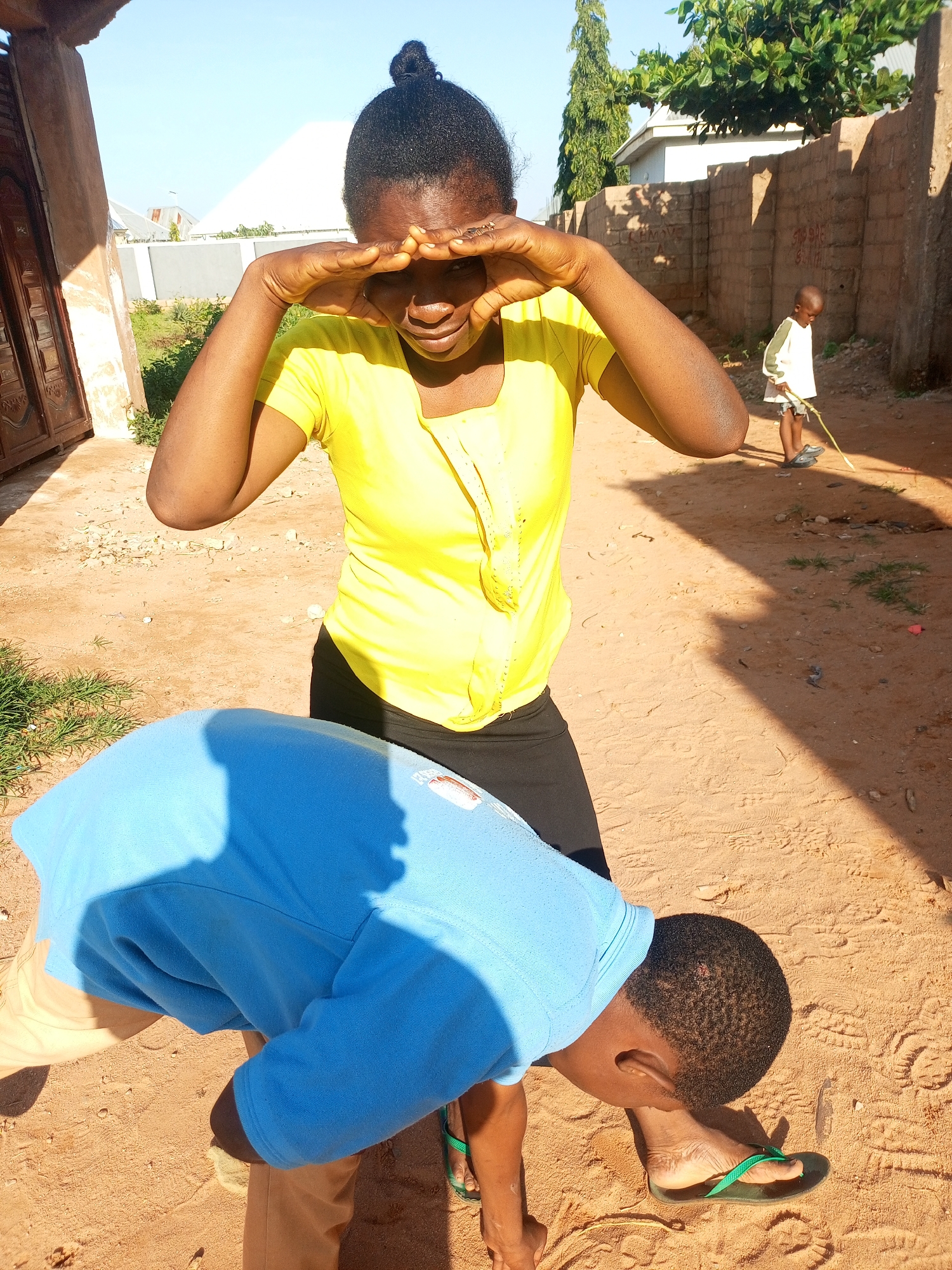 In the city of Tempure, Nigeria, a young Black female in a yellow t-shirt and black skirt stands in a sandy outdoor area, making a heart shape with her hands over her eyes. To her left, a young Black male wearing a blue sweatshirt and khaki pants bends over, his head down as if examining something on the ground. In the background, a young Black male in a white shirt and shorts stands with a stick. The environment consists of a sandy path, a brick wall, and some greenery, with a house visible on the left. The scene is illuminated by bright sunlight.