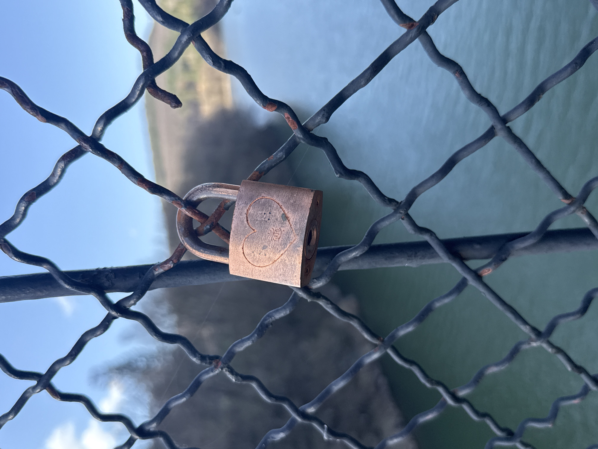 A close-up, eye-level shot shows a copper-colored padlock attached to a rusted metal wire fence. The padlock features an engraved heart design. Behind the fence, out of focus, is a body of water and greenery. The location is Dolni Pasarel, Bulgaria.