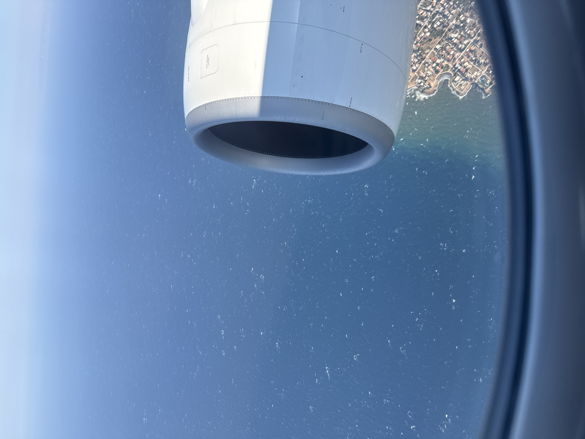 An aerial shot taken from an airplane window shows a vast expanse of the blue sea on the left and a coastal city on the right. The prominent object on the right side of the frame is the engine of the airplane, a large, white, cylindrical structure with a dark opening in the center. Text such as "NO STEP" and "P272 SENSOR ACCESS" is visible on the engine casing. Below the engine and on the right side of the image, the coastal city of Rafina, Greece, is visible. The city is characterized by a dense collection of buildings with red-tiled roofs, nestled along a curving coastline. Waves gently lap at the shore, and the sea is dotted with small whitecaps, suggesting a light breeze. The horizon is a clear blue, merging with the sky, indicating a bright, sunny day. The window frame of the airplane is visible at the bottom of the image, framing the aerial view.