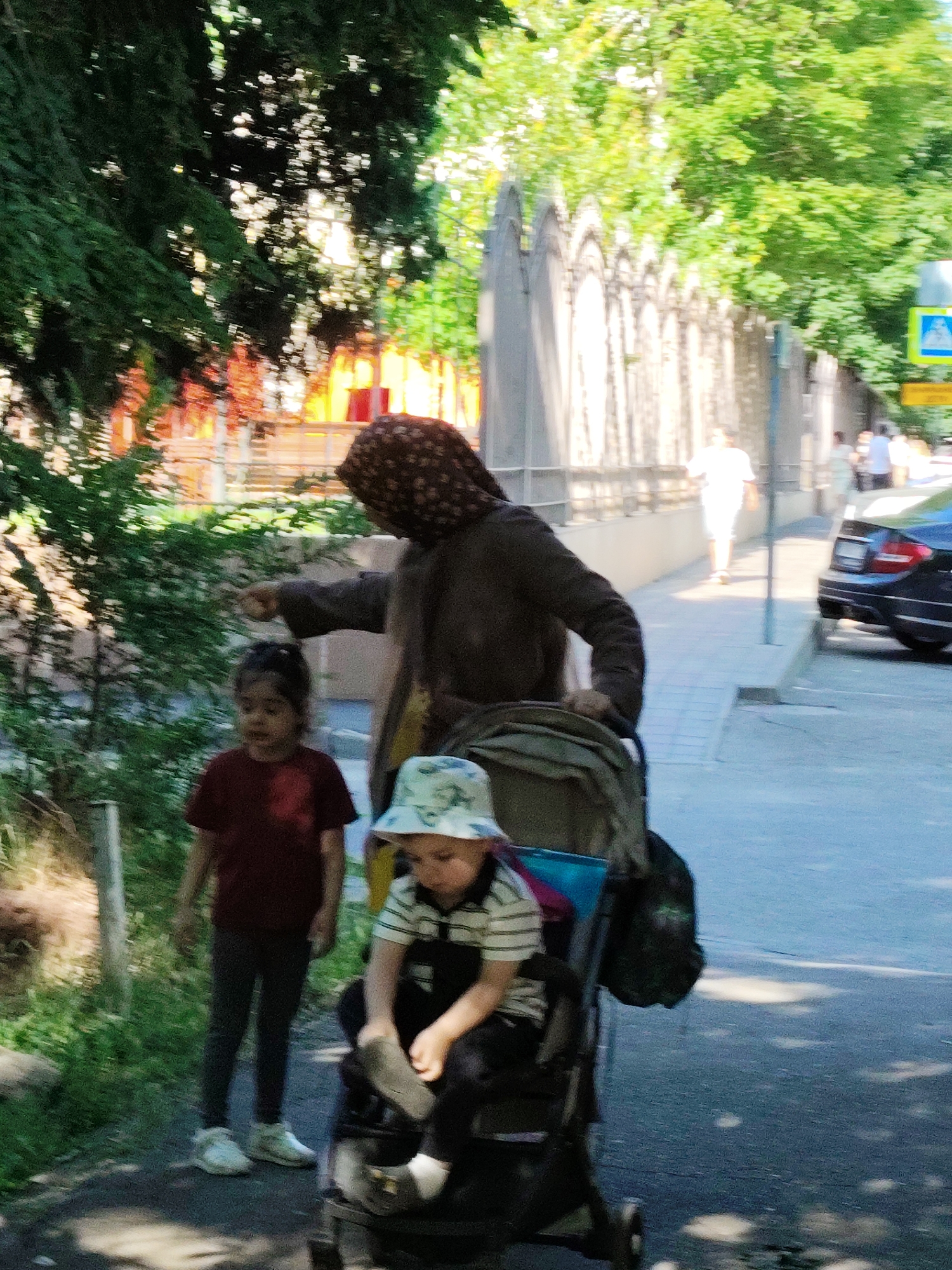 A partially blurred, sunny daytime exterior scene in a city shows a woman in a brown headscarf and dark coat, pushing a stroller containing a child wearing a patterned hat and striped shirt. A younger child in a red shirt and dark leggings stands beside the stroller, looking towards the woman. The woman is pointing with her right hand, possibly interacting with the younger child. The background features trees with green foliage, a decorative stone fence, a paved walkway, and a street with parked cars. A blue traffic sign with a white pedestrian symbol is visible in the upper right corner of the image. The overall impression is a candid moment of a family or group of people outdoors in an urban setting. While the specific location within Krasnodar, Russia, cannot be definitively determined from the image alone, elements like the architecture and signage are consistent with an urban environment in Russia.