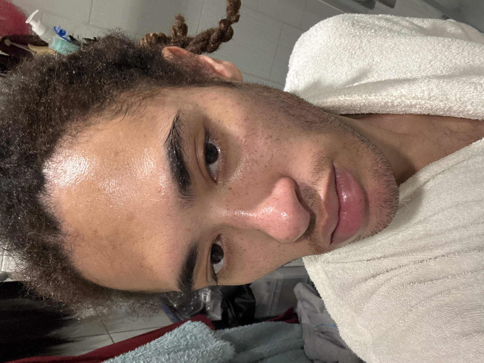 The image presents a close-up, head-and-shoulders portrait of an individual in an indoor setting located in Les Ulis, France. The subject is an adult male with medium-brown skin, characterized by visible freckles on the nose and cheeks, and a light stubble on the lower face. His dark, curly hair shows some areas styled into twists or dreadlocks on the right side of his head. The forehead and nose exhibit a shiny, wet appearance. He possesses prominent dark eyebrows and dark eyes, directed forward. He is wearing a white terrycloth bathrobe, visible around his neck and shoulders. The background on the left features a light blue towel draped, with obscured darker fabrics below. On the right, a light grey wall is visible, behind which a white pump bottle and a teal brush are discernable, along with other indistinct objects.