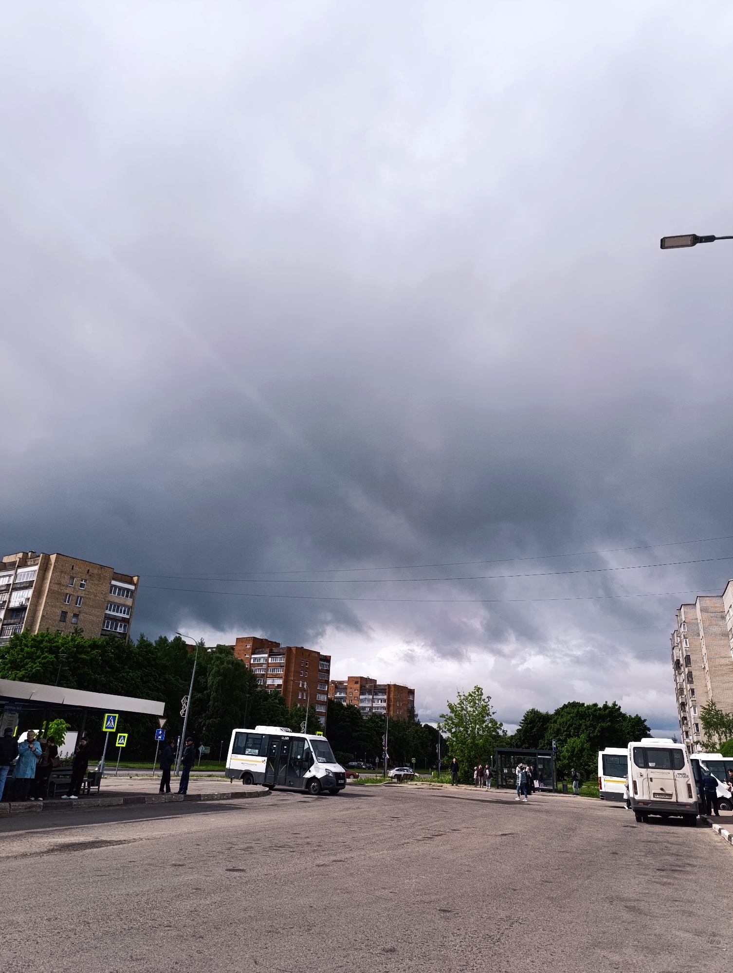 The image depicts a street scene in Chernogolovka, Russia, under a heavily overcast sky. Dark, ominous clouds dominate the sky, suggesting an impending storm.
Several buildings, likely apartment blocks, are visible in the background, constructed of brick and concrete.
In the foreground, an asphalt road stretches across the frame. A bus stop is situated on the left side of the road, with a few people waiting underneath the shelter. Pedestrian crossing signs are nearby.
Two white minibuses or shuttle vans are parked near the bus stop. People are standing and possibly interacting near these vehicles.
Electrical wires stretch across the frame, adding to the urban feel. A streetlight is visible on the right side of the image.