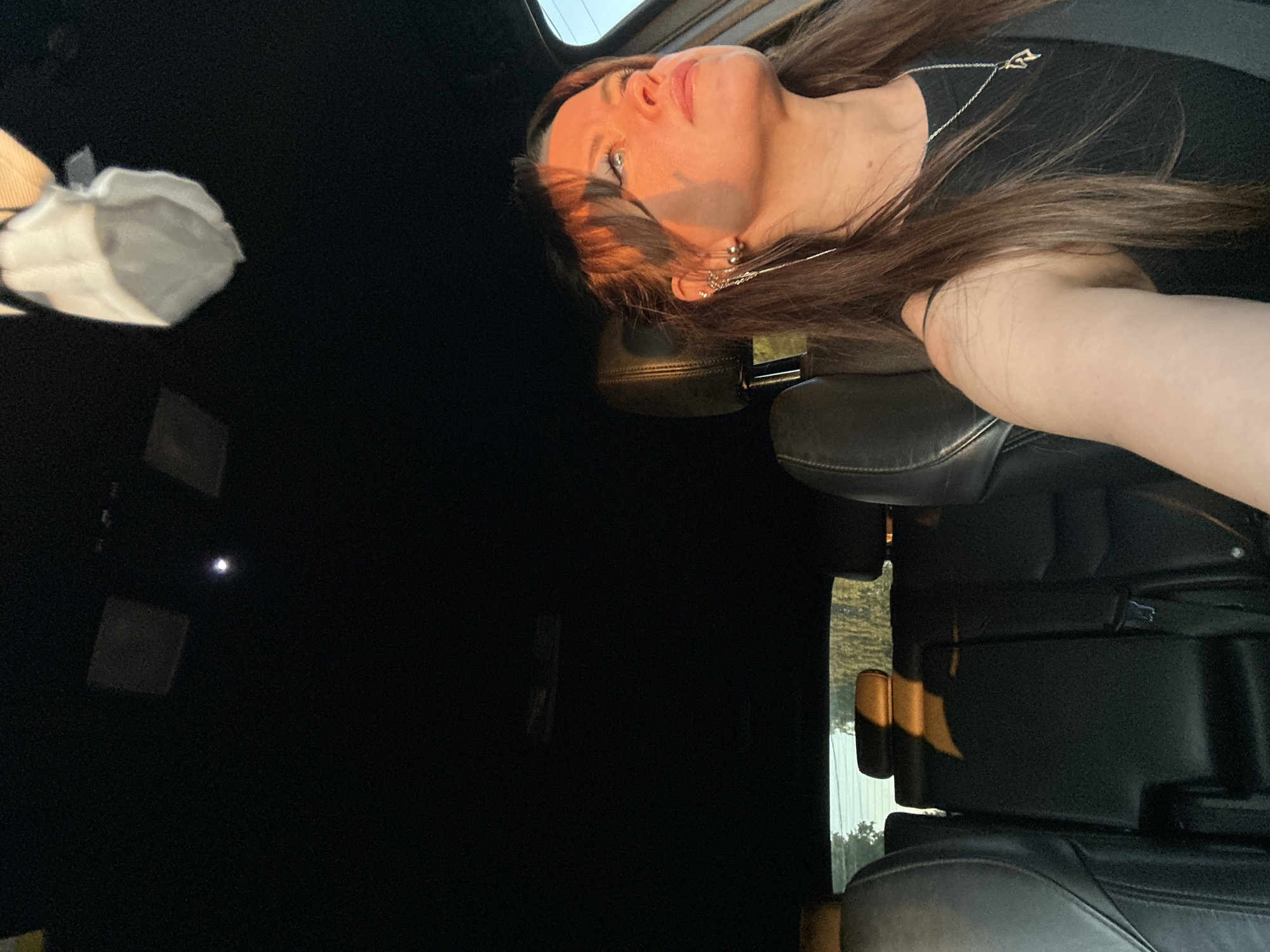 A woman is driving a car at sunset. The woman is wearing a black tank top and a silver necklace. She has long brown hair and is wearing a silver earring. The sun is setting, casting a warm glow on her face. The interior of the car is dark, with black leather seats. The back seats of the car are visible, as well as the headrests. The woman's face is illuminated by the setting sun. The car appears to be moving on a road in Chaikovsky, Russia, as indicated by the trees and foliage visible outside the car windows.