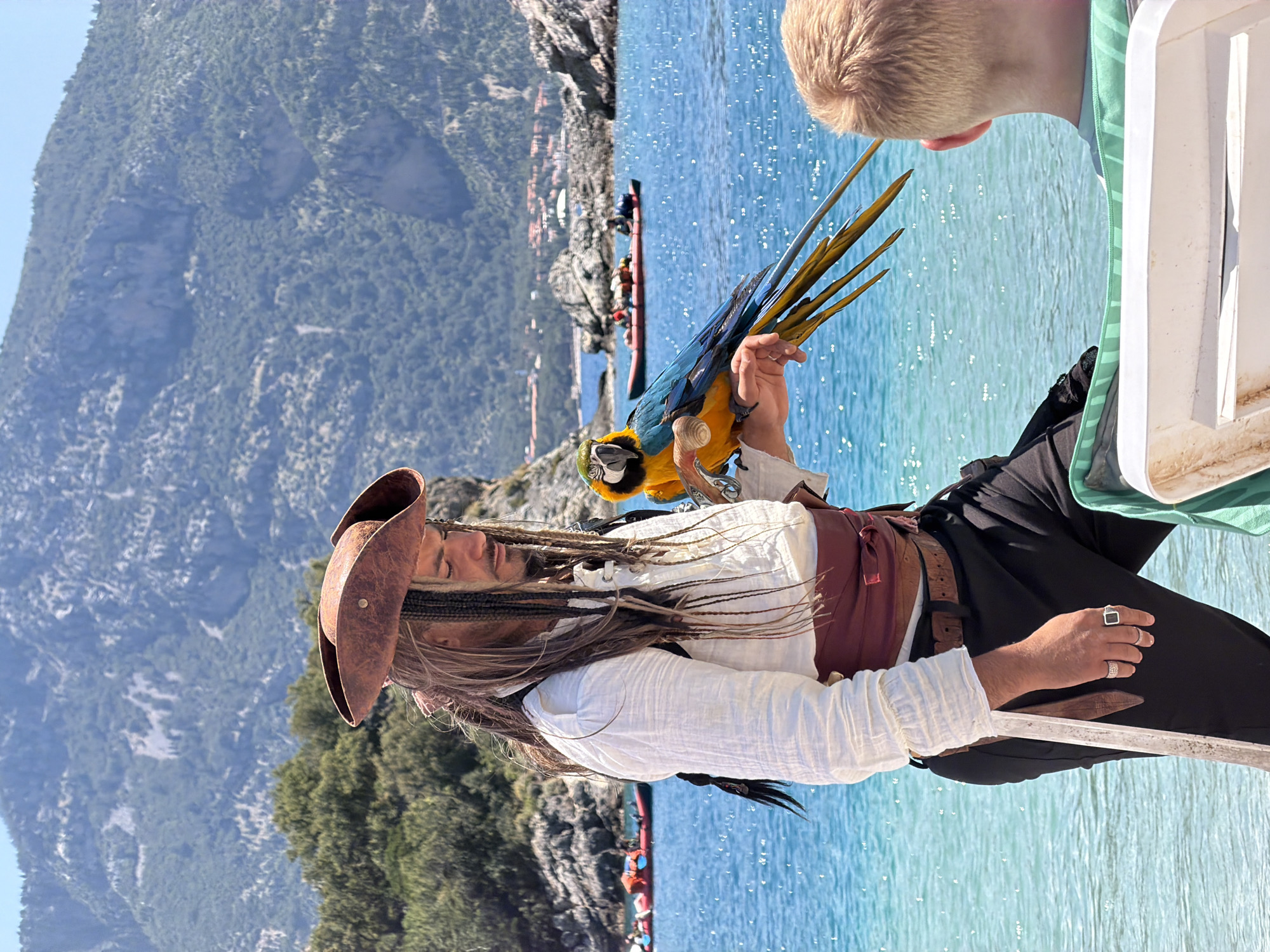A medium shot depicts a man dressed as Captain Jack Sparrow, holding a blue and yellow macaw parrot on his left arm. He wears a brown tricorn hat, white shirt, brown vest, and black pants with a maroon sash. His long hair is styled with braids and beads, typical of the character. He's wearing rings on his left hand and holding a sword. To his right, partially visible, is a child wearing a green and white top. The background features a body of water with a small boat carrying people. On the left, there is a forested area and a mountain range. The location is the city of Fethiye, Turkey, specifically near the sea or coastline, evident from the water and mountain range.