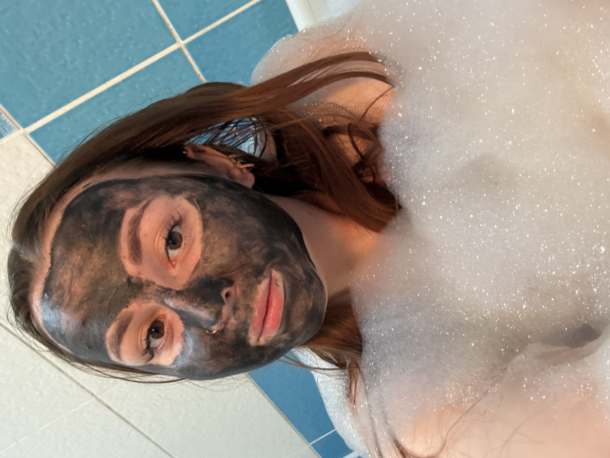 A young person, appearing female, is seen relaxing in a bathtub filled with a thick layer of white, glistening foam. The individual has long, dark brown hair spread out around their head and shoulders, partially submerged in the bubbles. Their face is covered with a dark, possibly charcoal, face mask, which leaves clear areas around the eyes and lips. The person's eyes are open and directed towards the camera, showing a neutral or slightly relaxed expression. A small silver nose piercing is visible on their right nostril. In the background, part of a tiled wall is visible, featuring alternating patterns of blue and white square tiles, indicating a bathroom setting. The overall scene suggests a moment of self-care or relaxation, with the person enjoying a bath and a beauty treatment. The image is captured from an elevated perspective, looking down at the subject. There is no visible text in the image. The specific time of day or precise location within Izhevsk, Russia, cannot be determined from the visual information provided.