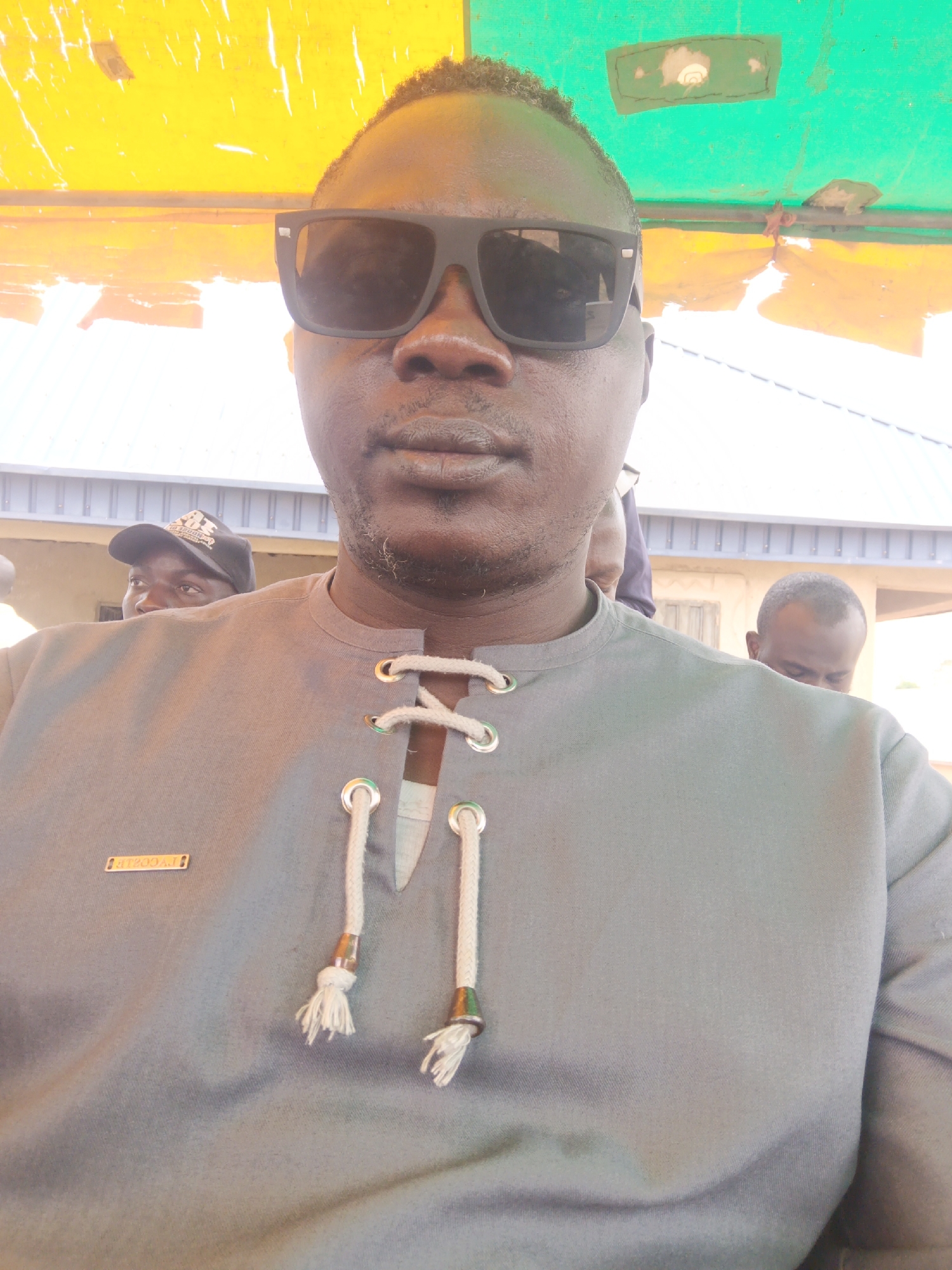 The image presents a medium shot of a dark-skinned man wearing large, dark sunglasses and a gray tunic-style shirt with a lace-up neck. The setting appears to be outdoors under a yellow and green awning or roof. Two other individuals are partially visible in the background. The location is Unknown, Unknown.