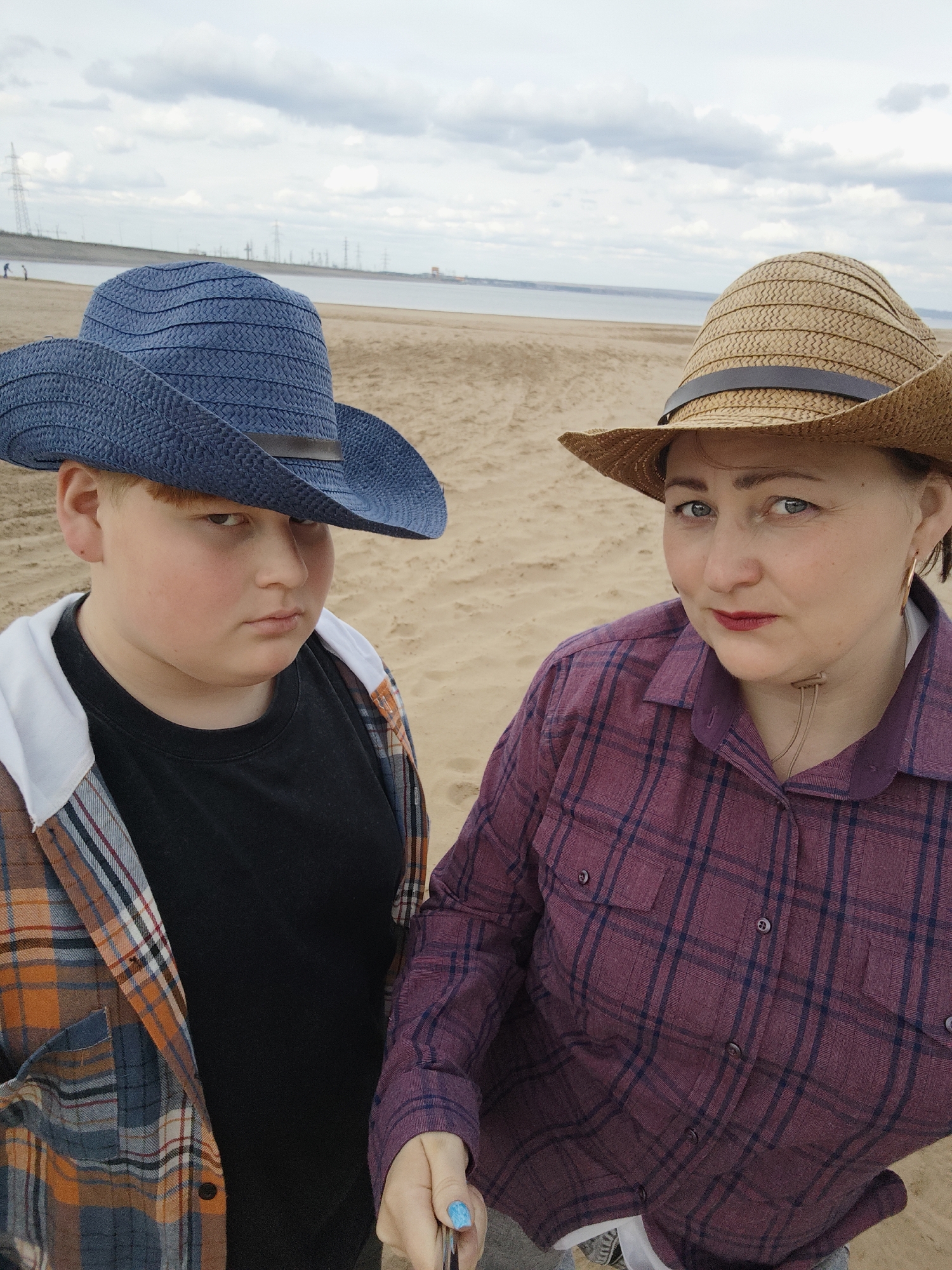 Two individuals, one appearing younger and the other older, are captured in a close-up shot outdoors. The younger person, to the left, wears a blue woven cowboy hat, a black shirt, and a multi-colored plaid jacket with a white hood visible. The older person, to the right, sports a straw cowboy hat, a plum-colored plaid shirt, and is holding what appears to be a selfie stick. Both are looking directly at the camera with serious expressions.
The setting is a sandy beach, with a body of water visible in the background. Beyond the water, infrastructure elements like power lines and industrial structures are discernible, indicating an urban or industrial backdrop. Based on the presence of industrial structures, water reservoir, and geographical factors, it is likely the location is the beach of Noviy, Russia. The sky is overcast with cloud coverage. The light is diffused.