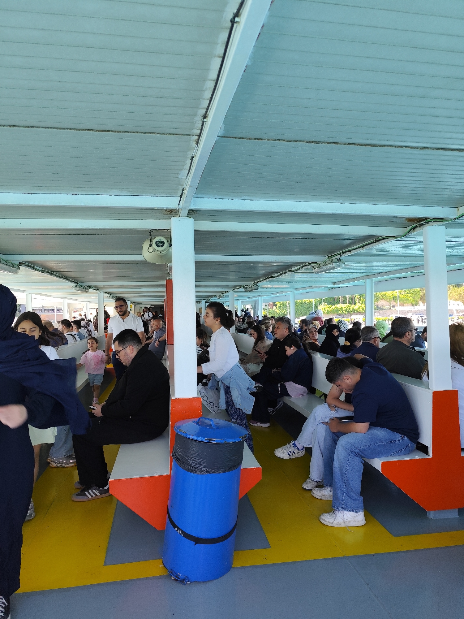 The image captures the interior of a ferry in Sarıyer, Turkey. Many people of varying ages sit on benches running lengthwise along the ferry's passenger area. A young girl walks down the aisle in the center. A blue trash can stands near the aisle. The ferry is covered with a light-colored roof, and sunlight filters through the open sides. Trees are seen in the distance through the ferry's open sides.
