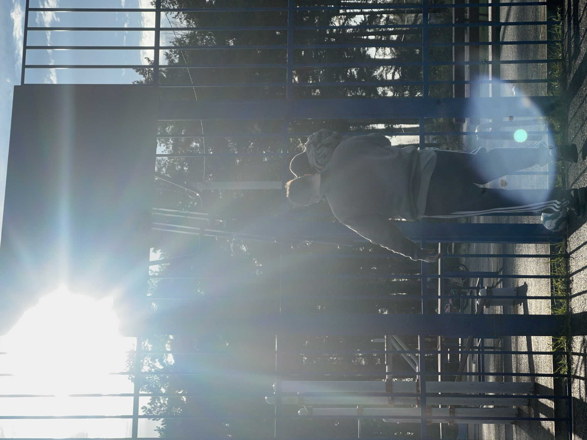 Here's a concise description of the image:
A person is visible through a blue metal fence. The person is wearing a gray sweatshirt and dark pants with light-colored stripes. The background behind the fence includes trees and sky. The presence of strong sunlight in the image makes it difficult to observe. The location is Раздолье, Russia.