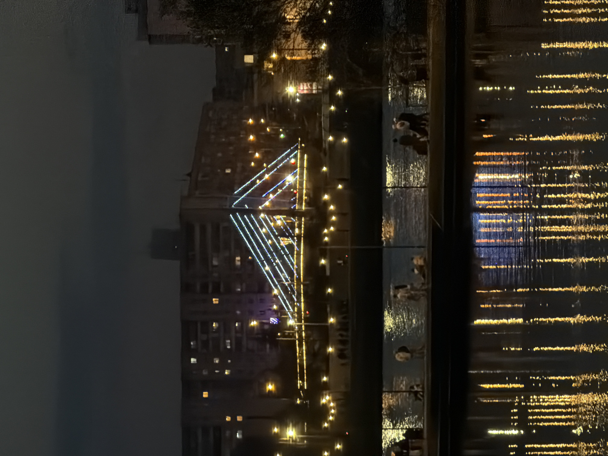 At night, a view of the Yenisey River in Krasnoyarsk, Russia. The river reflects the city lights, creating a shimmering effect. A lighted structure resembling a bridge or architectural display is prominent. Pedestrians are seen walking along the riverbank. The sky is dark and overcast. A multi-story building is visible in the background.