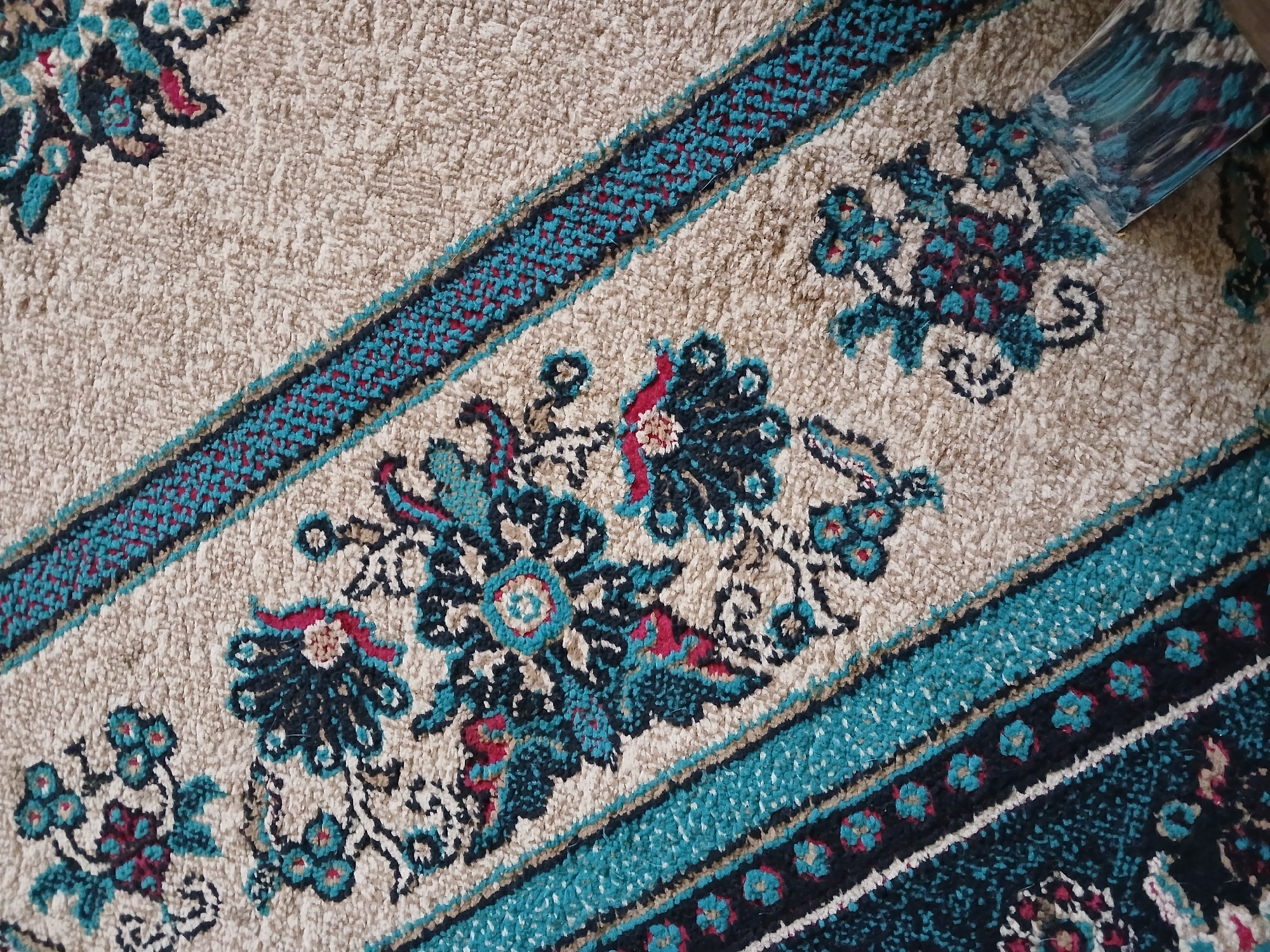 The image shows a close-up view of a carpet with an intricate floral pattern. The carpet has a predominantly beige background with diagonal bands of alternating dark blue and turquoise woven with red accents. The floral motifs are stylized, featuring dark blue and teal petals with hints of red and pink. The texture of the carpet appears plush and textured. In the bottom right corner, a portion of a cleaning tool, possibly a carpet cleaner attachment, is visible, suggesting an action of cleaning or maintenance. The scene is indoors, indicated by the lighting and the material of the carpet. The image itself does not provide any specific information about its location in the city of Guba, Azerbaijan, or any details about people or specific events.