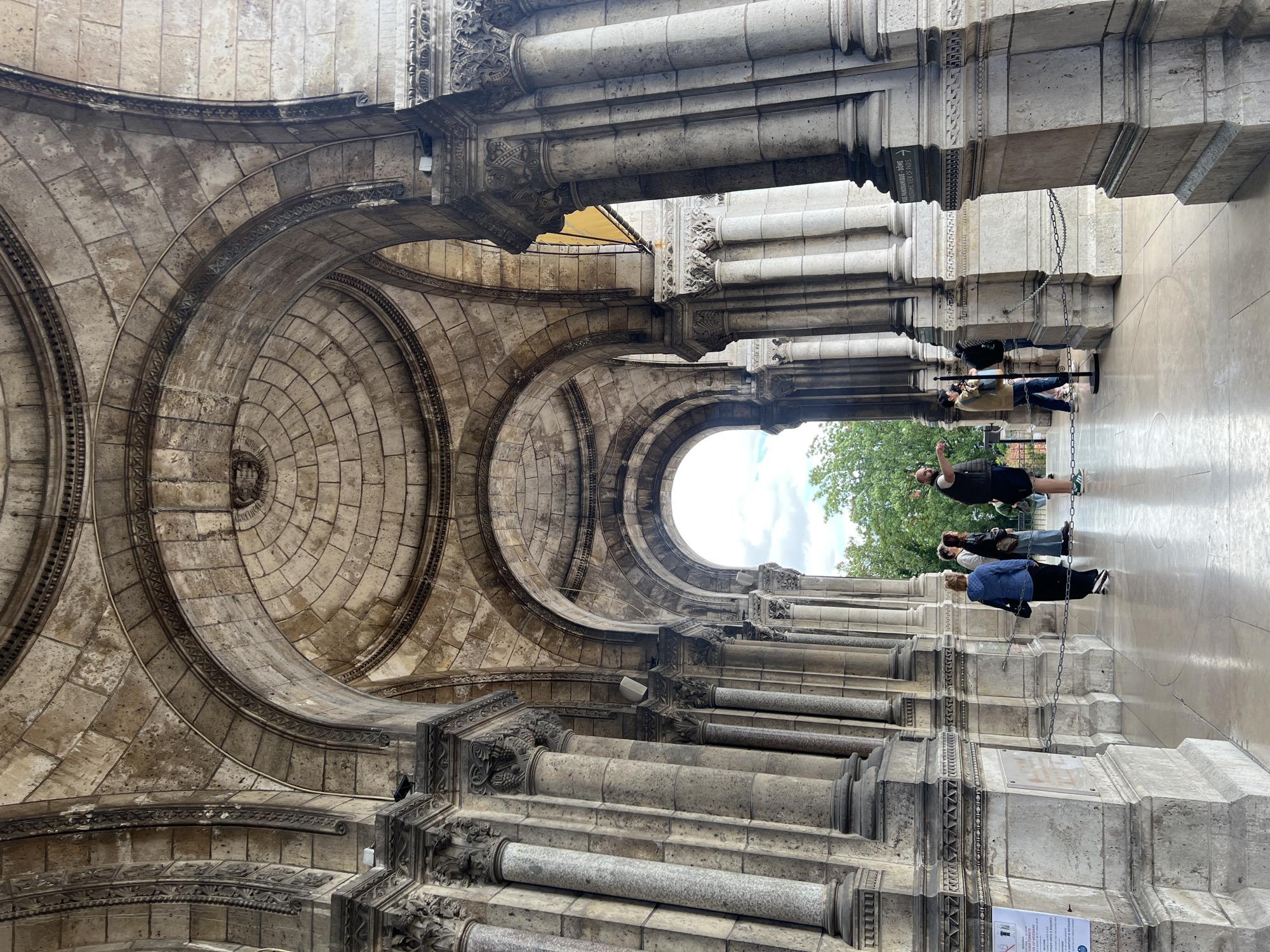 This low-angle photograph, taken with the camera rotated 90 degrees clockwise, captures the majestic stone architecture of a grand building, likely a basilica or cathedral. The upper portion of the corrected image showcases an impressive vaulted ceiling featuring intricate concentric stone arches and finely detailed carvings.
Towards the right side of the frame, several massive columns with ornate capitals create an open arcade, leading the eye towards an outside view. Bright, cloudy skies and lush green trees are visible through an archway, indicating an exterior setting. People are present in the lower right section of the image, standing between the columns. Some appear to be looking outwards or observing their surroundings; one individual in a dark vest is seen gesturing with their hand. Black chains are strung between some of the columns, possibly for crowd control or safety.
The architecture is characterized by its scale and intricate detail, with weathered light-colored stone suggesting age and historical significance. A sign attached to one of the columns is visible, bearing the text "VUE PANORAMIQUE-DOME" and "PANORAMIC VIEW OF PARIS," explicitly identifying the location as offering a scenic vista of the French capital. The time of day appears to be daytime, with soft, diffused light suggesting an overcast or partly cloudy sky.