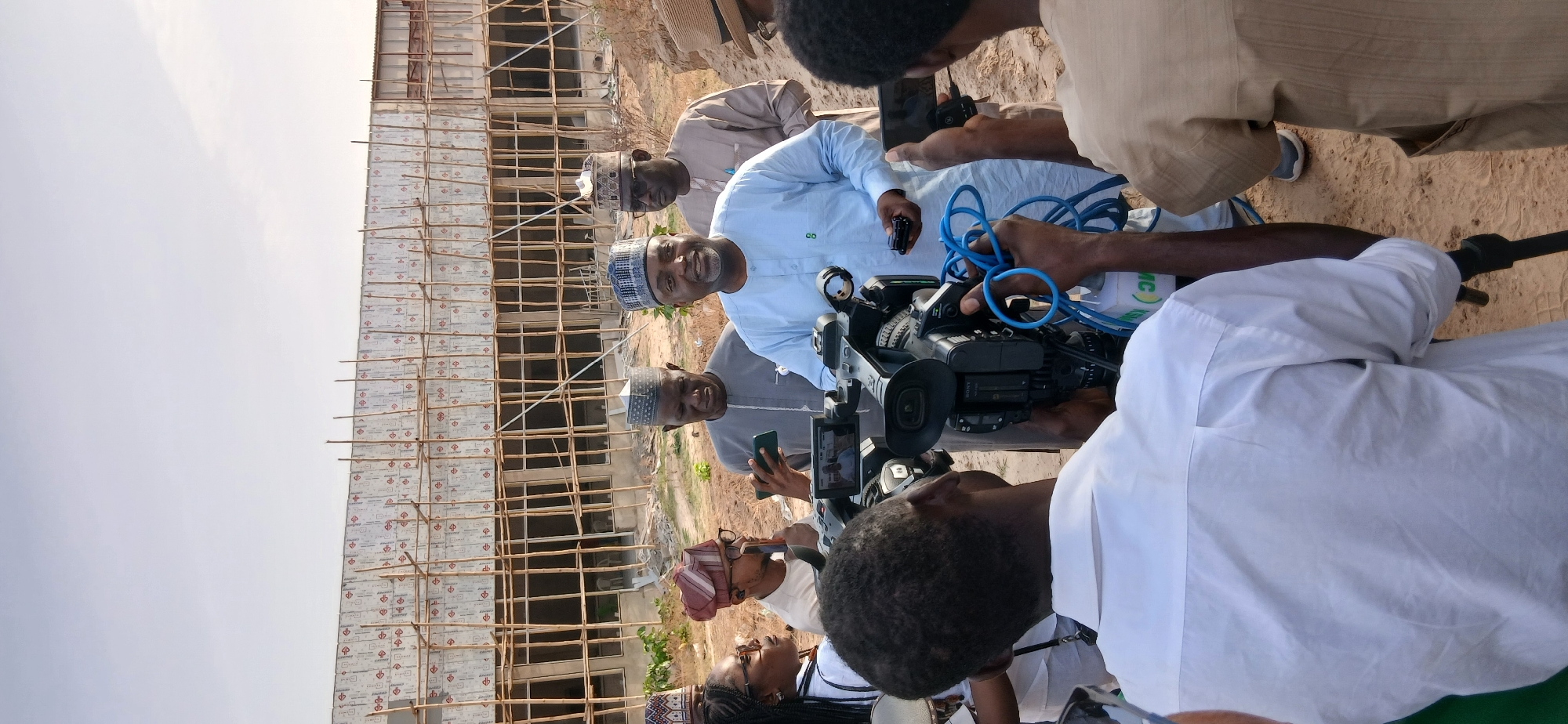 The image depicts an outdoor media event, likely a news report or interview, taking place at a construction site during daylight hours under a bright, clear sky.
Several individuals are present, with the focus on a group of men gathered around a professional video camera. A prominent figure, dressed in a light blue traditional kaftan and a patterned cap, is smiling warmly towards the camera, holding a small, dark object in his left hand. Another man in a light grey traditional outfit and cap stands behind him, also looking in the direction of the camera. A third man in a dark grey traditional garment and cap is seen to the left, holding a smartphone.
In the foreground, a camera operator's hands are visible, manipulating a black professional video camera, identifiable by "SONY" branding visible on its screen/body. A bright blue coiled cable is attached to the camera. Another person in a white top is partially visible in the immediate foreground, next to the camera. Further back, two individuals are seen from the side, one wearing glasses and a patterned headwrap.
The background features a multi-story building under construction, characterized by extensive wooden scaffolding. The building's exterior is covered in a white or silver material imprinted with small, repetitive red symbols or logos that are not clearly decipherable. The ground throughout the scene is sandy and appears to be bare earth. A white container with green text, including the letters "MC," is partially visible near the camera.