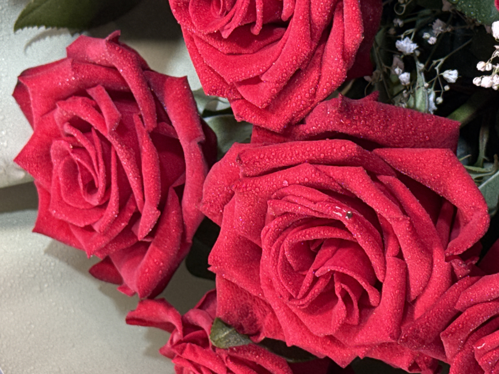 A close-up, overhead shot shows a bouquet of several red roses. The roses are deep red with many petals, and each petal is covered in tiny water droplets that glitter under the light. The background is blurred and appears to be a light gray surface with subtle texture, possibly a wall or fabric. Some small white flowers and green leaves are visible in the lower right portion of the image, adding a touch of contrast to the dominant red of the roses. The overall impression is one of freshness and natural beauty.