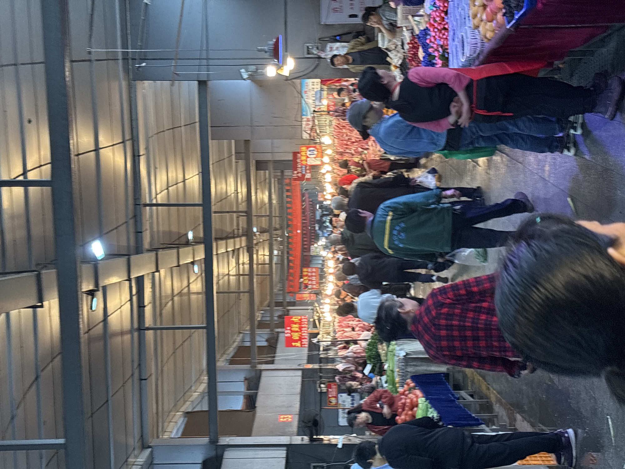 The image depicts a bustling indoor market scene in Jiangjin, China. Numerous people are browsing stalls filled with various goods, including produce, meat, and other items. The market is illuminated by overhead lights that reflect off the metal ceiling structure. People are interacting with vendors and examining merchandise. The environment is crowded and active, indicative of a typical marketplace.