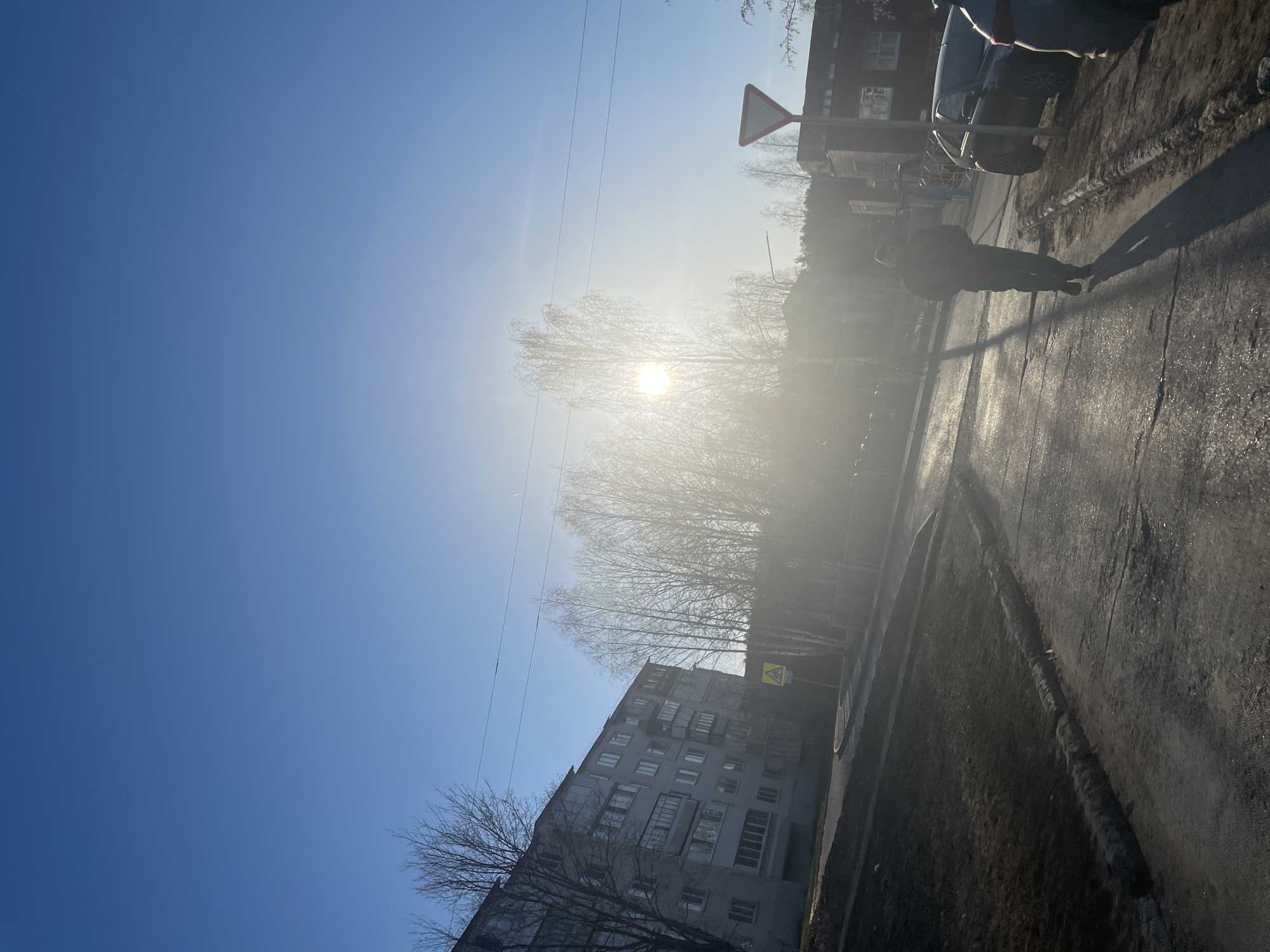 The media file displays an outdoor urban scene, captured with the camera rotated 90 degrees counter-clockwise. When viewed upright, the image depicts a clear, sunny day in what appears to be a residential area.
The sky, occupying the upper left portion of the frame (when upright), is a bright, cloudless blue. The sun, a brilliant white orb, is prominently positioned in the upper-middle, casting a significant glare across the scene. This strong, low-angle light produces long, distinct shadows, suggesting either early morning or late afternoon.
In the mid-ground, a lone individual is seen walking away from the camera on a paved surface, their figure largely silhouetted against the bright light. Their long shadow stretches out in front of them across the reflective road or sidewalk, which shows patches of what might be mud, ice, or wetness. Behind the person, a dark-colored car is parked on the side of the road.
The background features several bare deciduous trees, indicative of late winter or early spring, their branches silhouetted against the bright sky. Multi-story residential buildings, characterized by their blocky, functional architecture, are visible on the right side of the frame, hinting at a Soviet-era urban environment common in Russia. Thin power lines traverse the upper sky.
Two distinct traffic signs are visible: an inverted white triangle with a red border (a "Give Way" sign) is positioned near the upper right, and a yellow diamond-shaped warning sign with a black symbol of children crossing (indicating a children's pedestrian crossing) is further down near one of the buildings. There is no visible text. The overall atmosphere is bright and slightly hazy due to the sun's glare, capturing an everyday moment in Noviy, Russia.