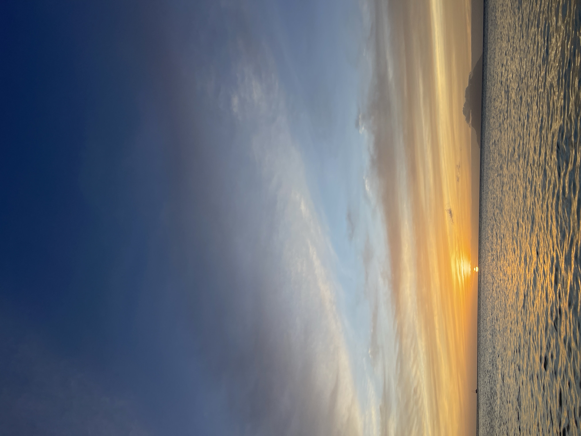 The image captures a serene sunset over the ocean in Mare Anglaise, Seychelles. The sky displays a gradient of colors, transitioning from deep blue at the top to lighter shades of blue, pink, and orange towards the horizon. Wispy clouds are scattered across the sky, catching the warm hues of the setting sun. The sun itself is a bright orange orb, just above the horizon, casting a golden reflection on the gently rippling water. In the distance, a small, dark silhouette of a boat is visible on the left, and a faint outline of a mountainous island is seen on the right. The water occupies the lower portion of the frame, reflecting the colors of the sky and the sun. The overall scene is one of tranquil natural beauty.