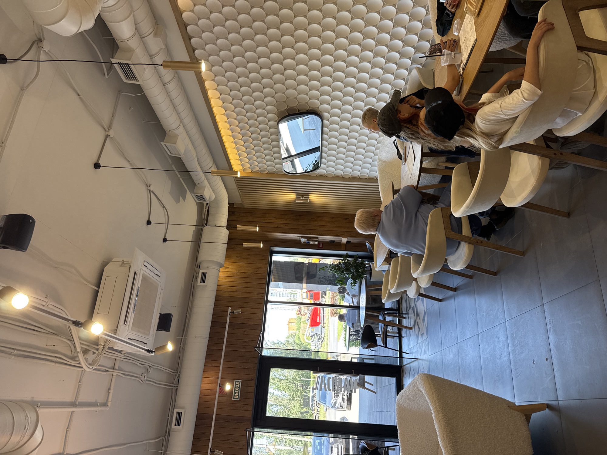 The image shows the interior of a cafe with several people seated at tables. The cafe has a modern design with a white, textured ceiling featuring circular patterns. Exposed white pipes run along the ceiling, and golden pendant lights hang down. Wooden accents are visible on the walls and in the furniture.
In the foreground, a person is seated at a table, their back to the camera, wearing a blue shirt. Several other individuals are visible, including a group at a table in the middle ground where a person in a white shirt is seated. Some people are wearing hats, and one person has blonde hair.
The cafe appears to be well-lit, with natural light coming from large windows and doors at the far end of the establishment. The weather outside seems sunny, indicated by the bright sky visible through the windows. The overall atmosphere appears casual and relaxed.
There is a visible air conditioning unit mounted on the ceiling. A sign with Russian text, "ВЫХОД" which means "EXIT", is visible on a wall near the entrance. The floor is tiled with a grey pattern. The activity taking place is people dining or socializing at the cafe.