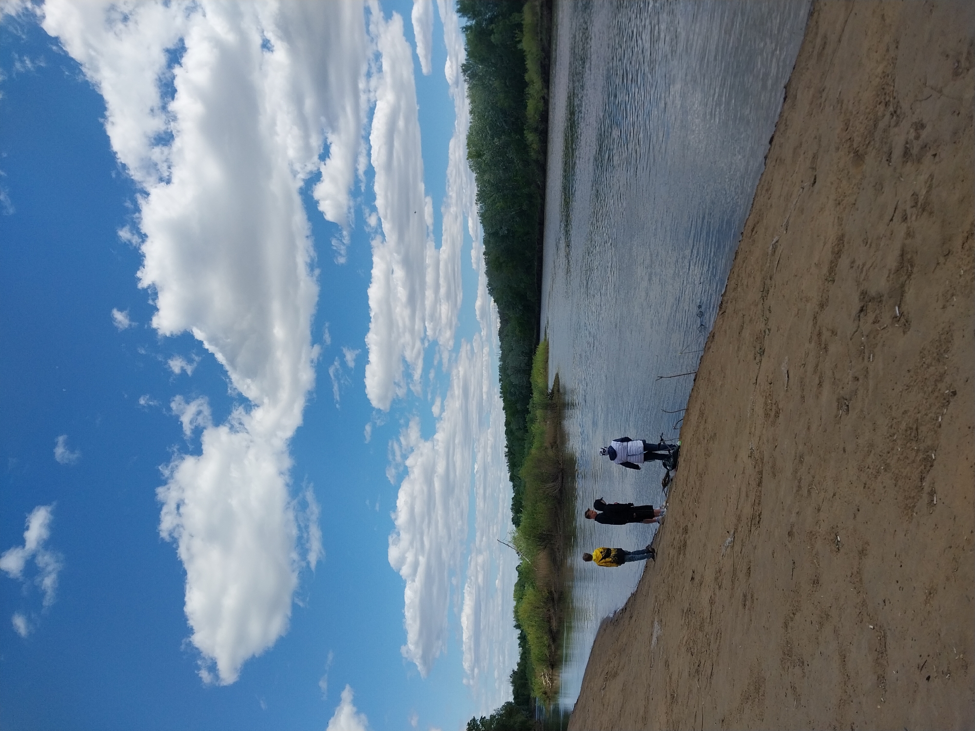 Three people are standing on a sandy riverbank beside a body of water. The person closest to the water is wearing a white jacket and standing near a bicycle. The second person is wearing a black jacket, and the third is wearing a yellow jacket. The riverbank is made of sand and slopes down towards the water. Across the river, there is a line of trees. The sky is blue with white clouds. It is daytime. The location is Orenburg, Russia.