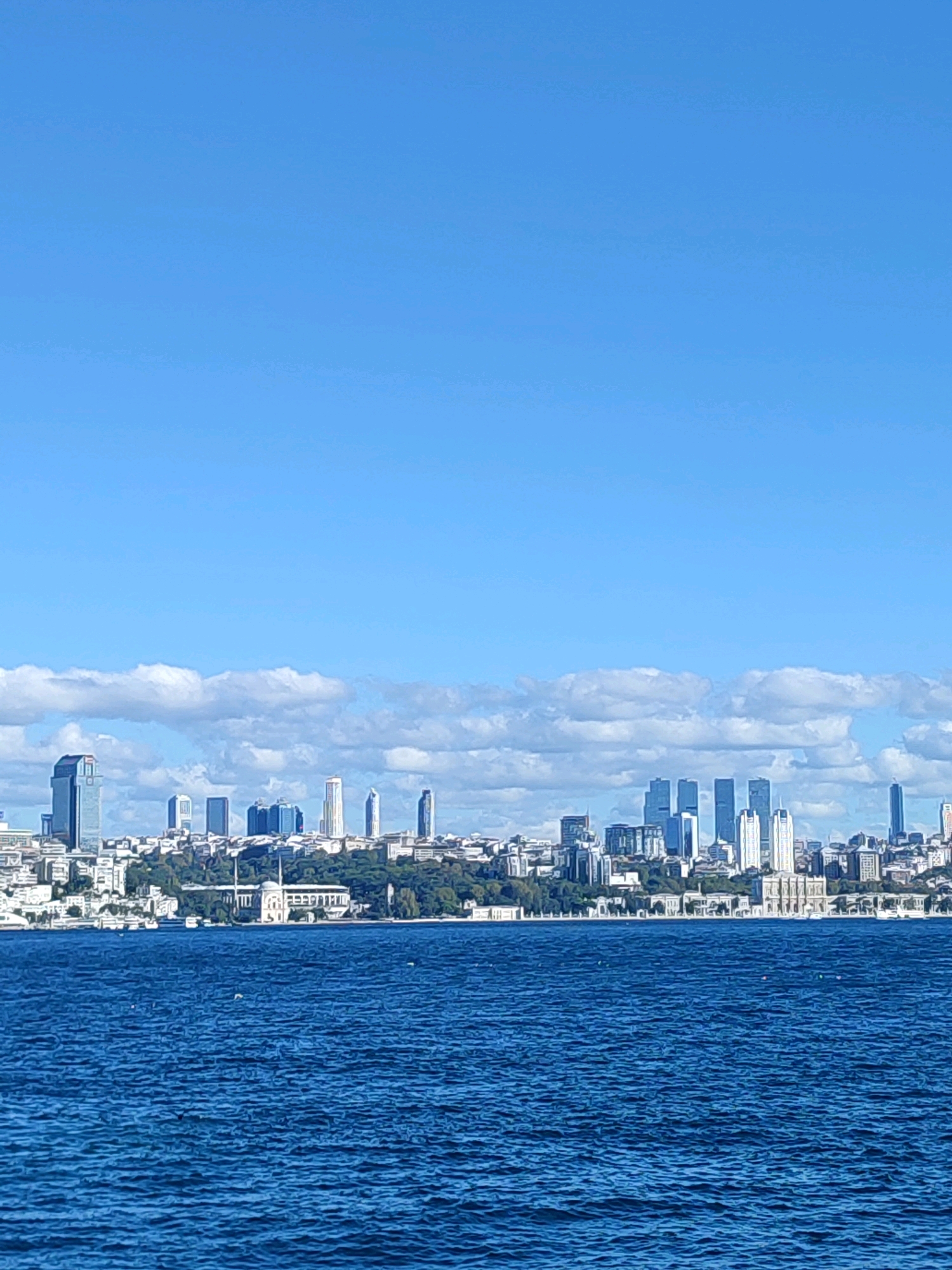 The image shows a panoramic view of the Üsküdar district in Istanbul, Turkey, from across the Bosphorus Strait. The foreground is dominated by the deep blue, slightly rippled water of the strait, with a few white birds visible on the surface. In the middle ground, the Üsküdar skyline rises, characterized by a mix of modern skyscrapers and older, white-washed buildings. A prominent structure with domes and minarets, likely a mosque complex, is visible towards the left of the mid-ground. The background features a clear blue sky with scattered white clouds. The overall scene depicts a bright, sunny day with the cityscape reflecting on the water.
