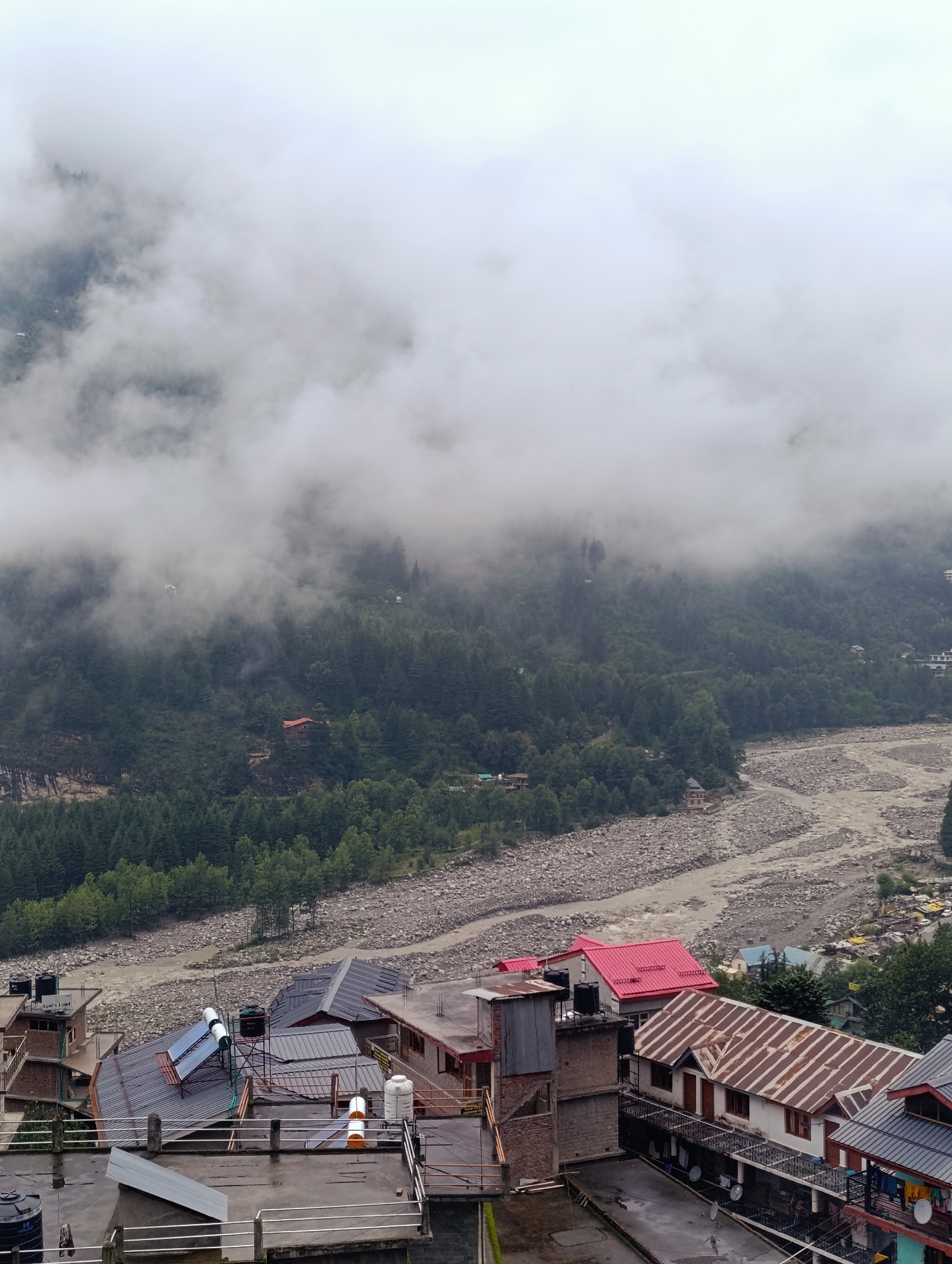 This is an aerial shot of the town of Vashisht, India, on a cloudy day. The foreground shows several buildings with various roof types including corrugated metal, asphalt shingles, and solar panels on a sloped roof. There are also water tanks visible on some of the rooftops. In the mid-ground, a wide, rocky river flows through a valley. The riverbed is filled with stones and boulders. On the banks of the river and the slopes of the valley, there are many trees forming dense green forests. In the background, mist and clouds obscure the mountain peaks and parts of the forest, giving the scene a dramatic and atmospheric feel. A small red-roofed building is visible nestled amongst the trees on the mountainside.