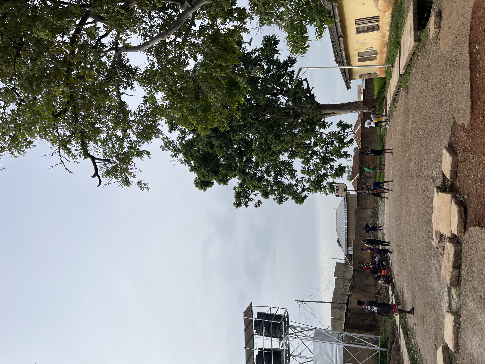 A group of young people are gathered in what appears to be an outdoor street or court area. Several individuals are playing basketball, with one person actively dribbling a ball, and others observing or waiting their turn. A basketball hoop is visible in the background. The scene is set outdoors, with a large tree providing shade. Buildings, including one with a yellow facade and boarded-up windows, are present in the background, suggesting a residential or community setting. The ground is a dirt path with some concrete slabs. A large water tank structure is also visible. The weather appears overcast. The overall atmosphere seems casual and engaged, with the focus on the basketball activity. The image is oriented in landscape format with the bottom of the frame on the right. There is some illegible text on a sign in the background. The time of day appears to be daytime, likely afternoon.