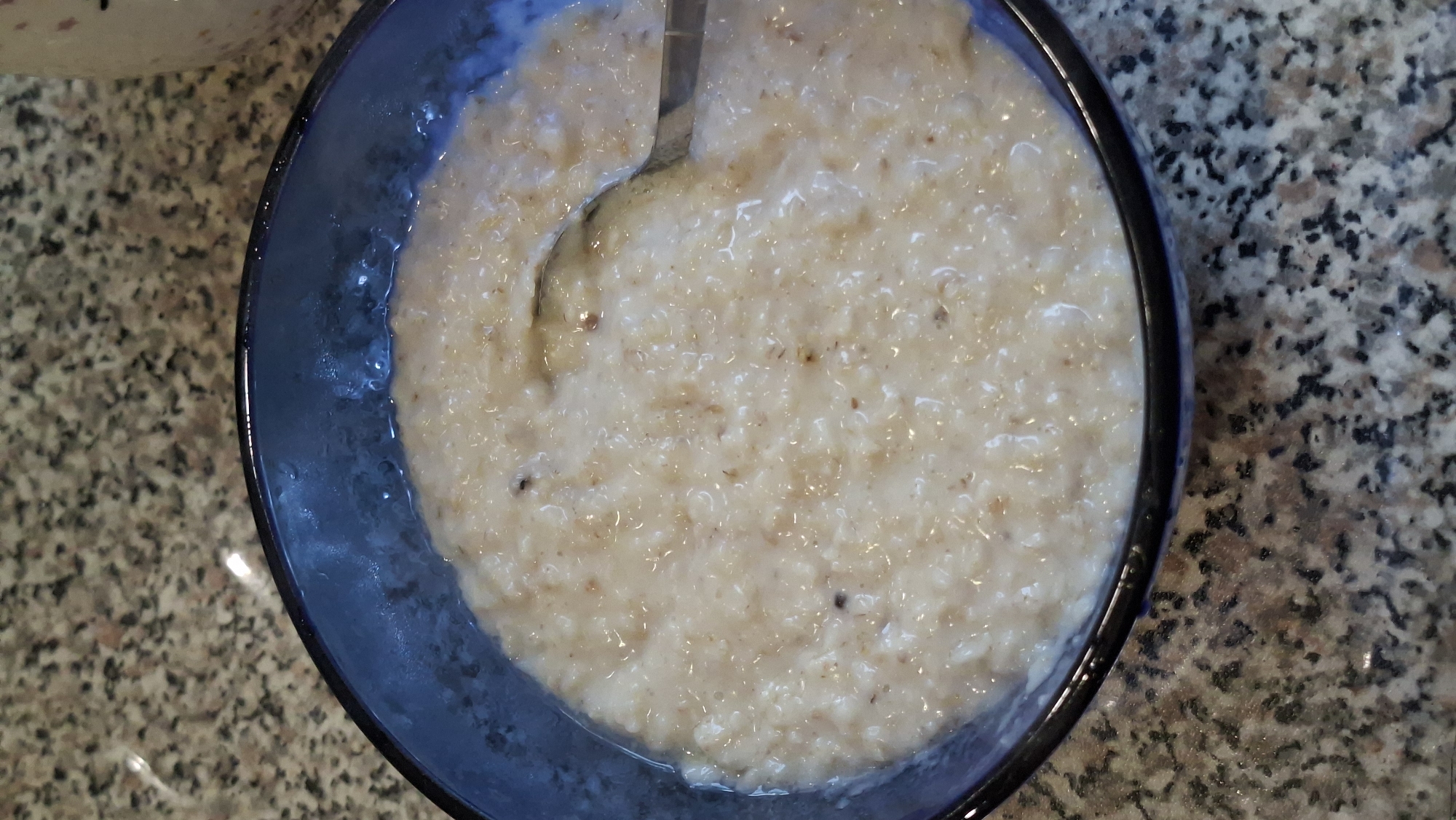 A close-up overhead shot shows a blue bowl filled with oatmeal. A spoon is partially submerged in the oatmeal, suggesting it is being eaten. The oatmeal is a light tan color and appears to have small brown flecks throughout. The bowl is placed on a speckled granite countertop. The background is blurred but shows a glimpse of what appears to be a granite surface and a white object. The image is well-lit and sharp. No specific location in Myadzyel, Belarus is visible.