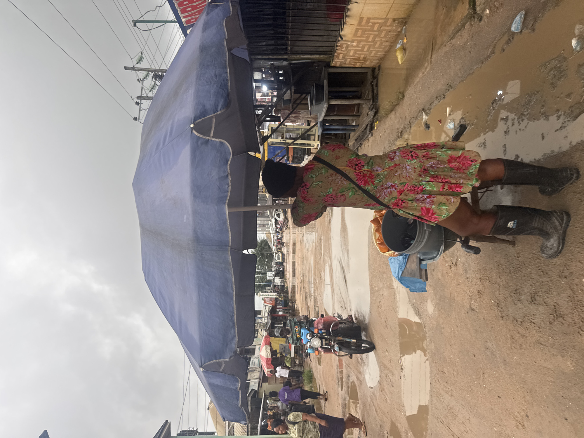 A woman wearing a floral dress and black rubber boots stands under a large blue umbrella in a flooded street in Ihiagwa, Nigeria. The street is unpaved and covered in mud and puddles, with debris scattered around. A motorcycle is visible in the background, ridden by a man wearing a green shirt and a cap. Other people and market stalls line the sides of the street. The sky is overcast and gray. The scene depicts a typical market day in a location prone to flooding.