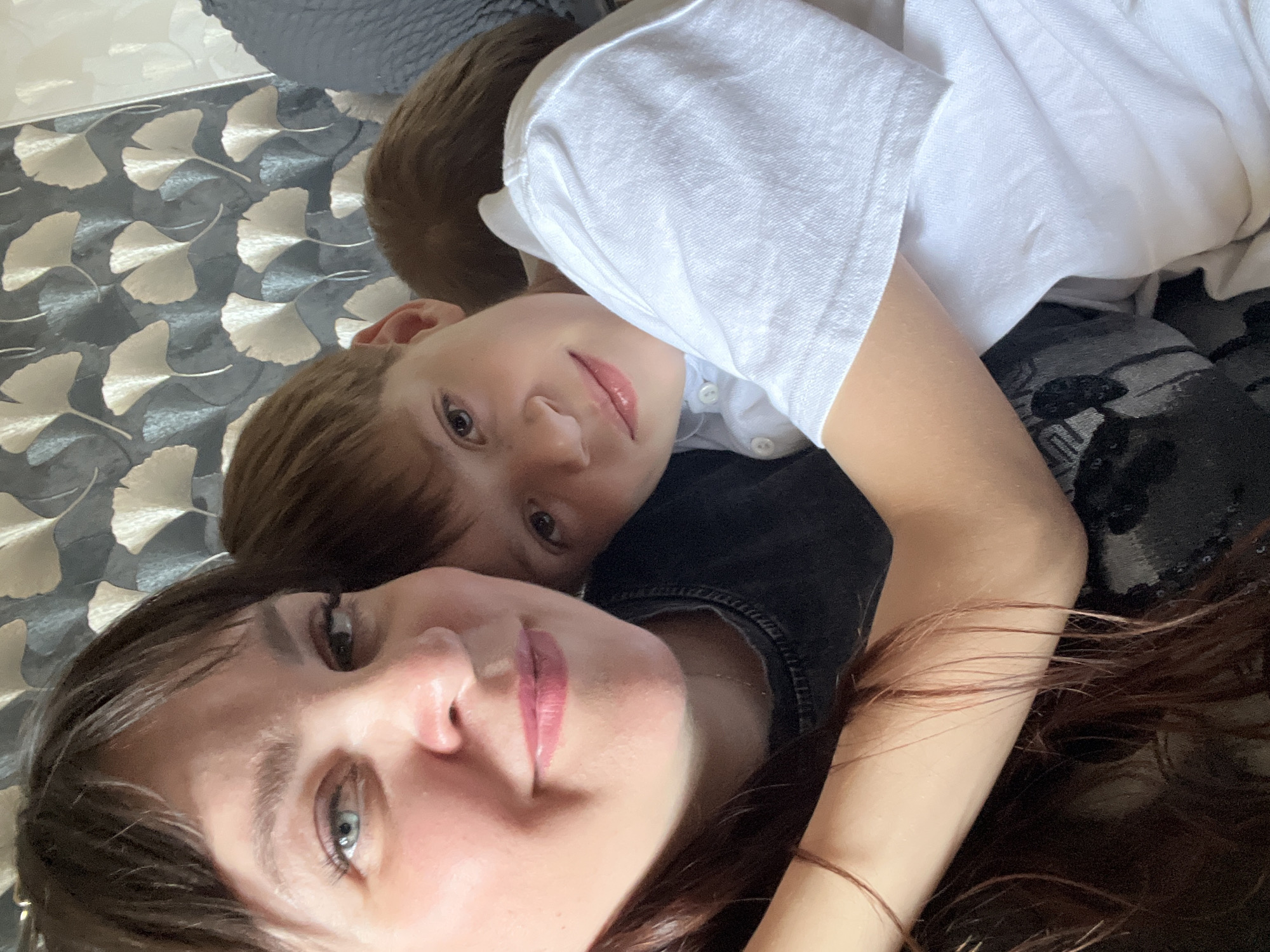 A selfie-style photograph shows a woman and a boy lying closely together. The woman, in the foreground, has fair skin, light blue eyes, and long brown hair. Her lips appear to be tinted pink. The boy, slightly behind her, has light brown hair and light-colored eyes. He wears a white t-shirt. The woman's dark shirt is visible underneath. The background appears to be a gray and white patterned surface, possibly a bedspread or wallpaper. The location is Chaikovsky, Russia.