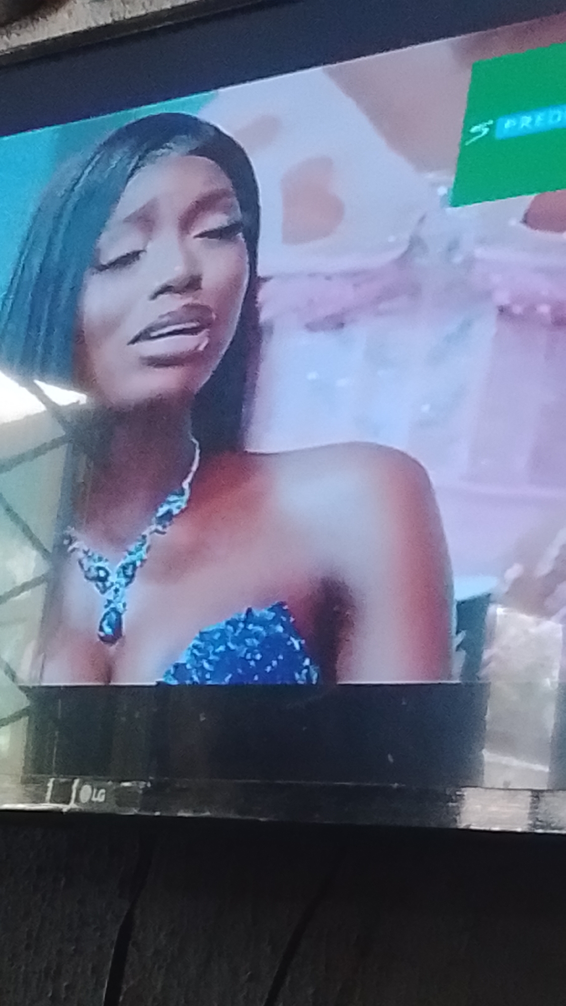 The image is a close-up, low-angle shot of a woman's upper body and face, captured from a television screen. The woman has dark skin, black hair styled straight, and is wearing heavy eye makeup and dark lipstick. She has a pronounced expression with her mouth slightly open. She is adorned with a sparkling blue sequined dress and a multi-faceted blue jeweled necklace. To the upper right of the frame, a green graphic with white text, possibly a logo or channel identification, is partially visible. The bottom edge of the television screen is visible, displaying an "LG" logo. The background is blurred and appears to be a pastel-colored, possibly celebratory, setting. The scene is not identifiable as being located in Lokoja, Nigeria.