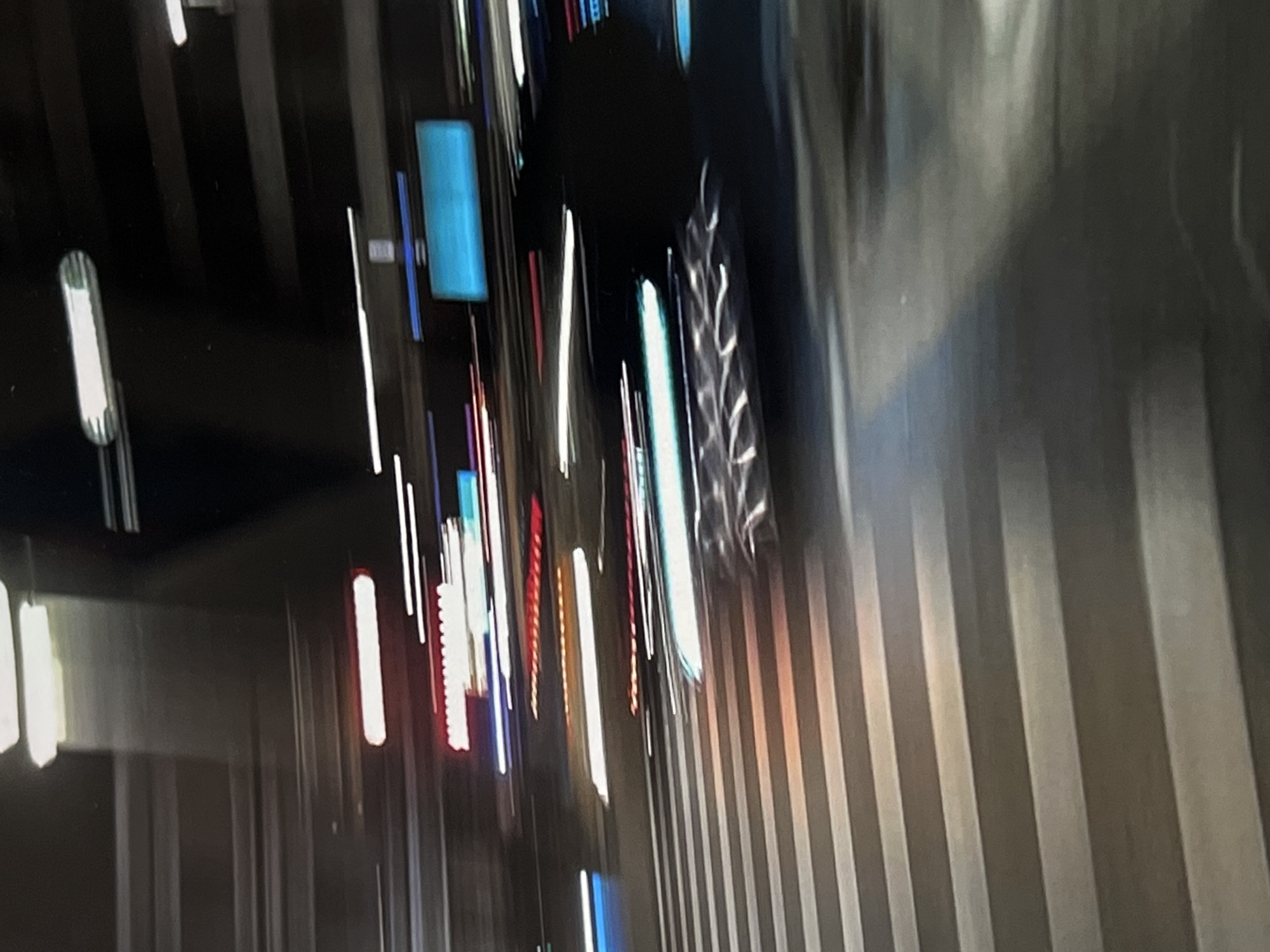 The image depicts a motion-blurred night scene in what appears to be an urban environment. Horizontal streaks of light, characteristic of moving vehicles or illuminated signage at night, dominate the composition. The foreground shows what seems to be a crosswalk with painted white lines, some illuminated by the vehicle lights. In the background, various colored lights, including white, red, and blue, are streaked horizontally, suggesting traffic and possibly advertising or building lights. The dark areas suggest a night sky or unlit portions of buildings and roads. The overall impression is of movement and the ephemeral nature of urban light at night. There is no specific visual evidence within the image to confirm its location in Genemuiden, Netherlands.
