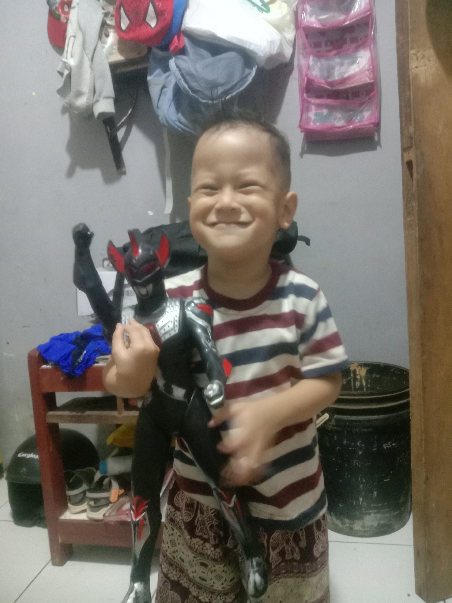 A young child with a wide smile holds a black and red toy action figure. The child is wearing a striped t-shirt and a patterned skirt or sarong. The action figure has a black body with red accents on its head and legs, and it is positioned in front of the child. The background appears to be an indoor setting, possibly a room in a home, with various items visible. To the left, there are some shelves with clothing and bags, including a red Spider-Man hat. A pink hanging organizer with multiple compartments is attached to the wall. To the right, there are large black cylindrical containers and a wooden doorframe. A red wooden shelf unit with shoes and a blue cloth is also visible in the lower left of the image. The lighting is somewhat dim, and the overall scene suggests a casual domestic environment. The location is identified as Cianjur, Indonesia.