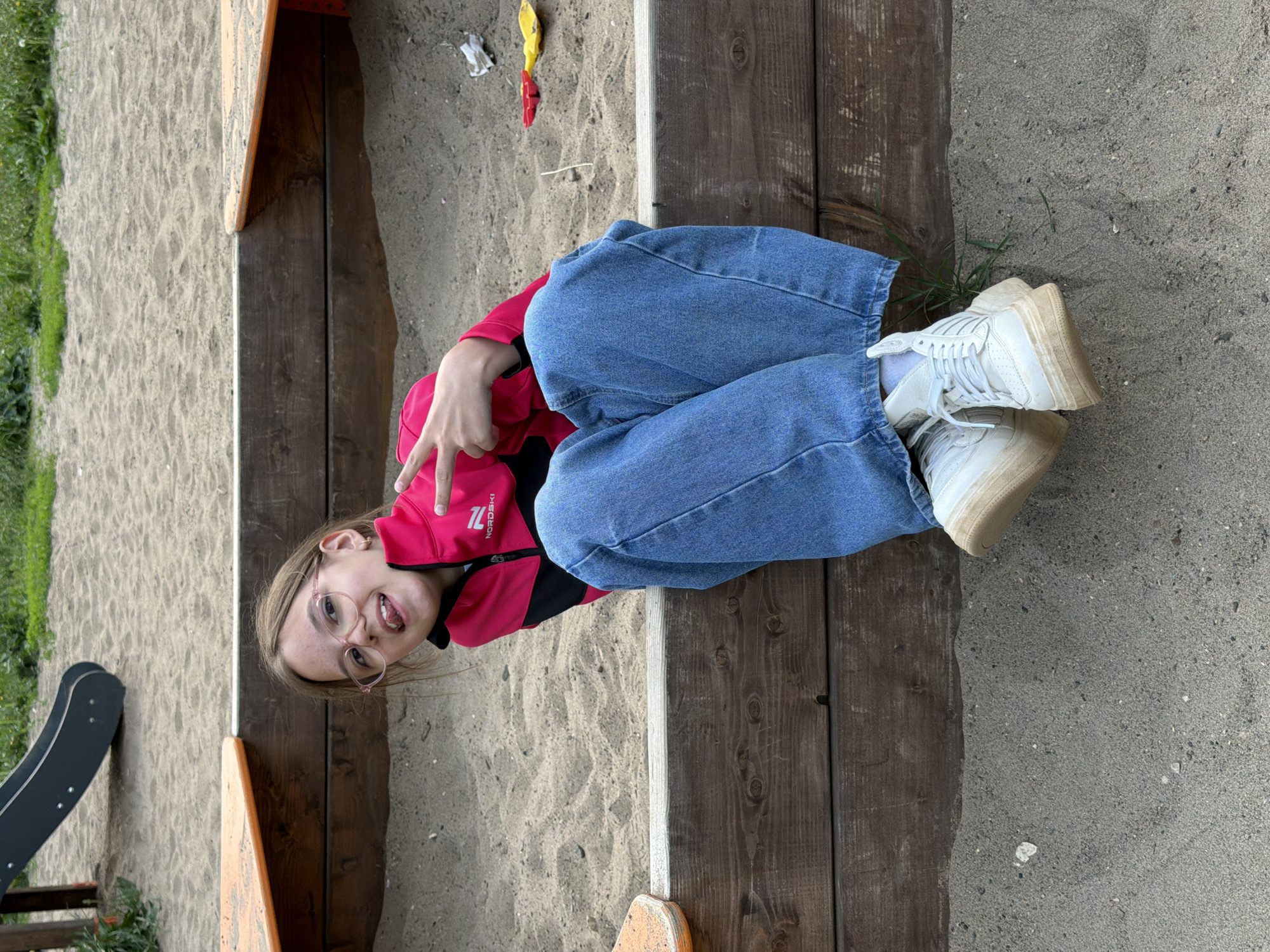 A young person is lying on a wooden edge of a sandbox, with blue jeans, a red and black jacket, and white sneakers. They are wearing glasses and making a peace sign with their hand. The sandbox is filled with sand, and there are some toys scattered around. There is a slide and grass visible in the background. The scene is in Chaikovsky, Russia.