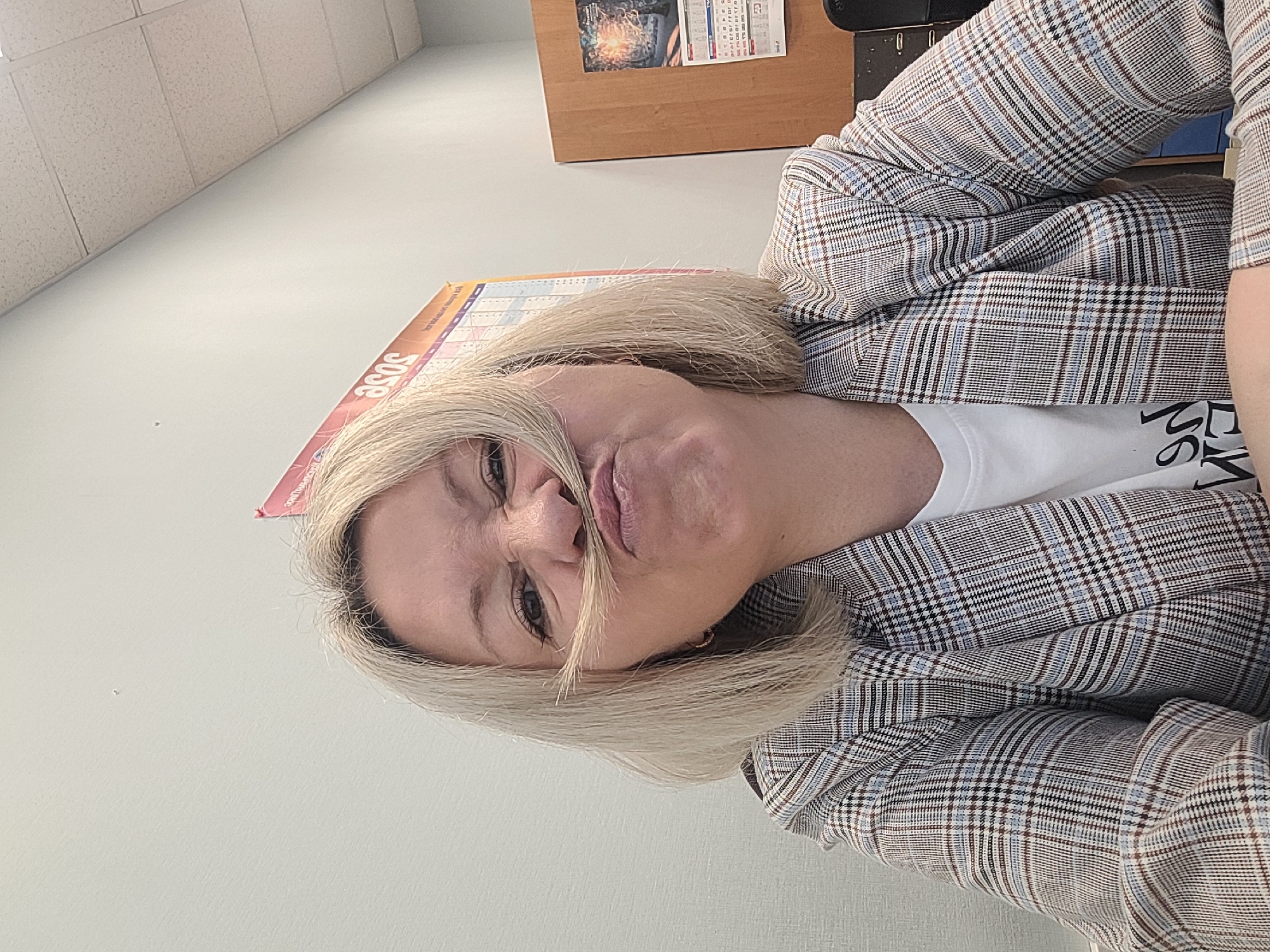 A woman with blonde hair is indoors, likely in an office, making a funny face. She has a strand of her hair arranged to resemble a mustache above her lip. She is wearing a white t-shirt and a plaid blazer. A calendar is visible in the background. The exact location in Izhevsk, Russia is not determinable from the given information.