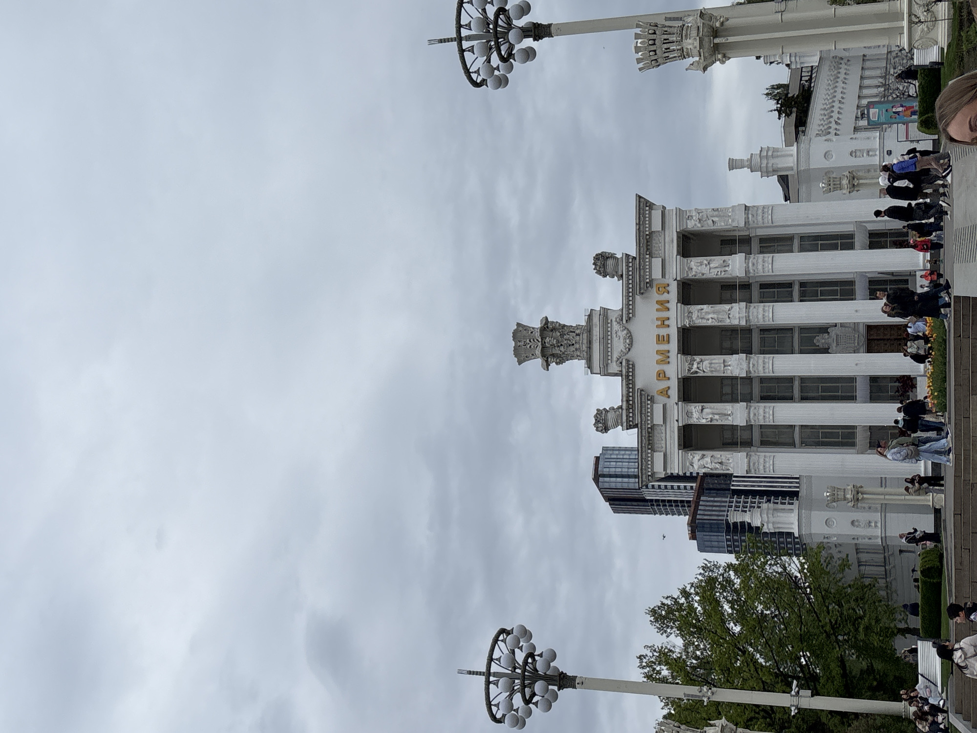 The image depicts the Armenia Pavilion at the Exhibition of Achievements of National Economy (VDNKh) in Moscow, Russia. The building is white with classical columns and golden lettering spelling "Армения" (Armenia) above the entrance. People are seen walking and sitting in front of the pavilion. Modern buildings are visible in the background, juxtaposing with the classic Soviet architecture. The sky is overcast. There are street lamps with multiple globe lights on tall poles surrounding the architecture.