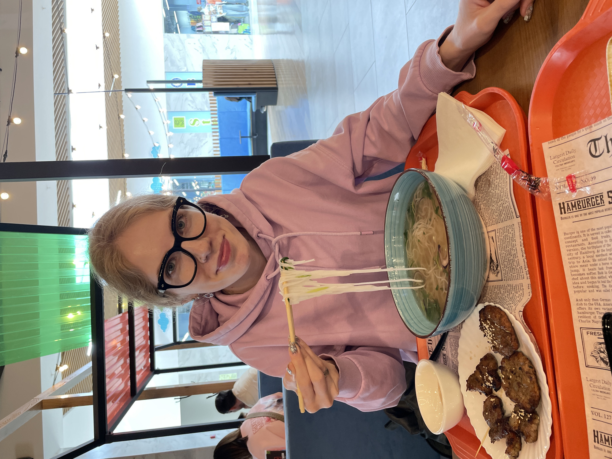 A woman is sitting at a table in a brightly lit indoor location, likely a food court or restaurant, potentially within a shopping center, identified by the text "Izhevsk Russia" being visible in the bottom right corner of the original, unedited image file. She is wearing a pink hooded sweatshirt and glasses. She is holding chopsticks with noodles and is about to eat a bowl of noodles. On the table is a tray with the bowl of noodles, a small dish with a dipping sauce, and a plate of grilled meat. A newspaper-printed placemat is visible under the dishes.