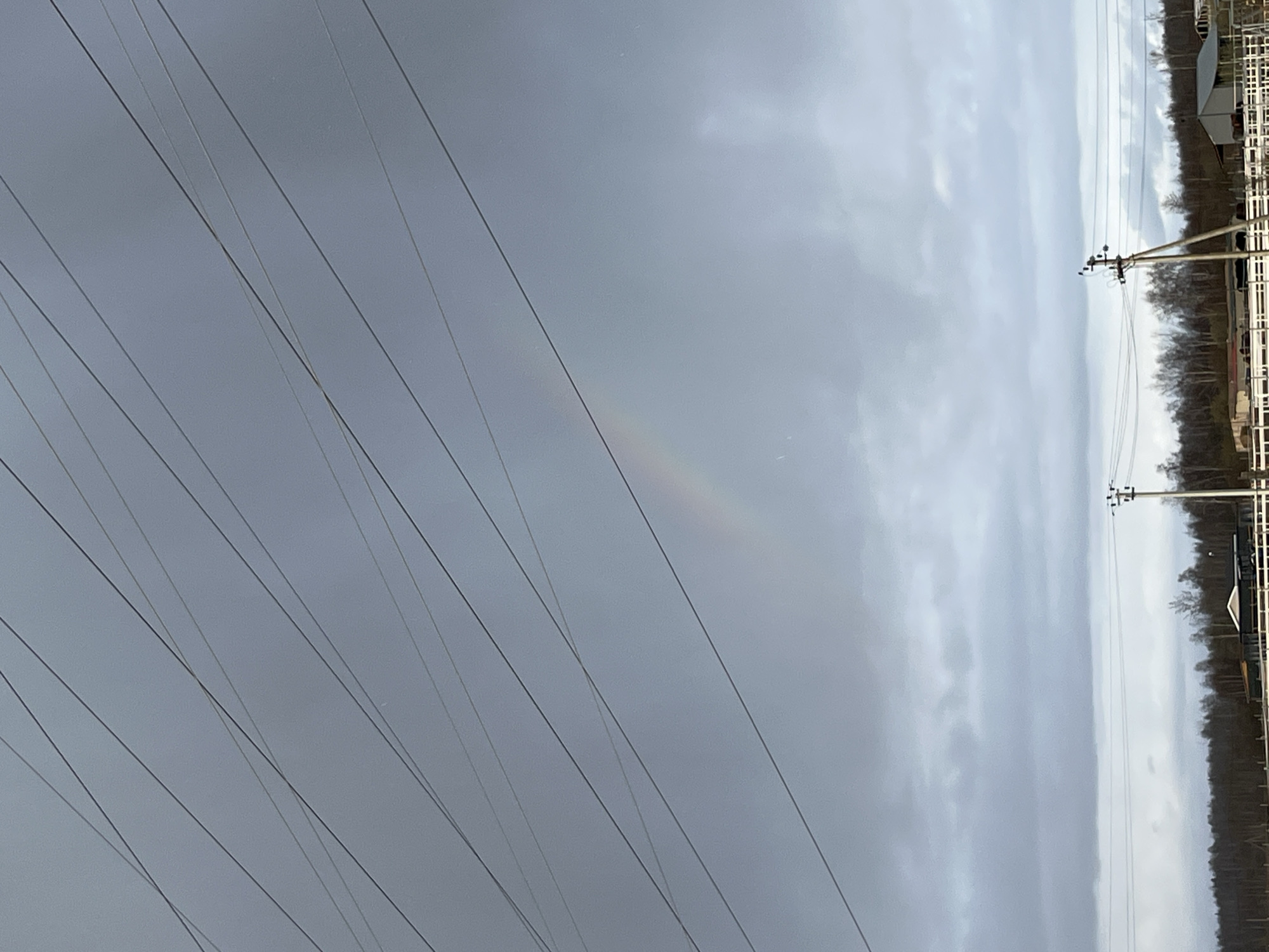 The image shows an overcast sky in Razdol'e, Russia. A faint rainbow is visible. Electrical power lines extend across the left side of the frame. On the right, there are wooden utility poles and the edge of a wooded area with what appears to be a white fence and some structures.