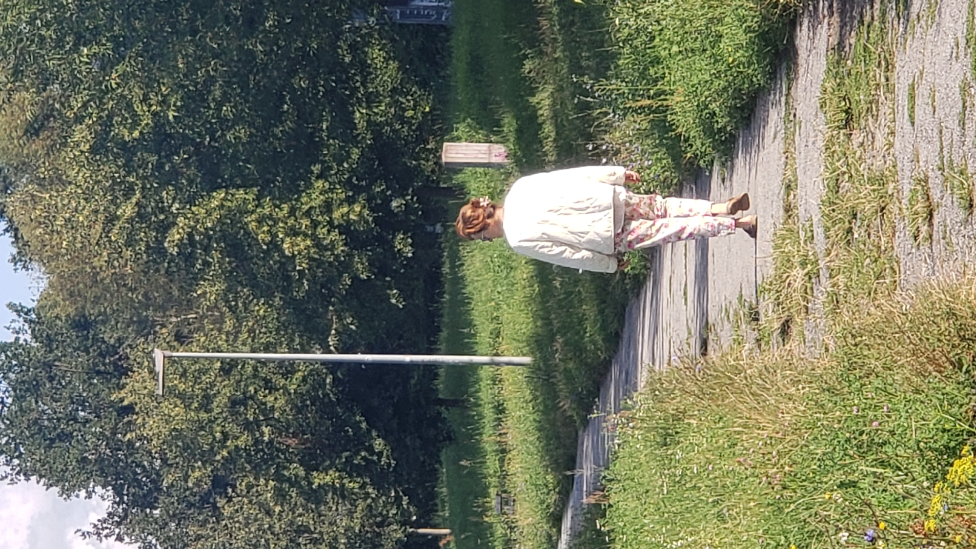A person with red hair pulled up in a bun and wearing glasses is walking away from the camera on a paved pathway. The person is wearing a light-colored quilted jacket and floral patterned pants. The pathway is bordered by grass and trees. A street light is visible on the left side of the pathway. In the background, there are more trees and some buildings that appear to be in disrepair. The scene suggests an urban or semi-urban environment, possibly a park or a neglected area within the city of Prešov, Slovakia. The lighting indicates it is daytime, with sunlight casting shadows.
