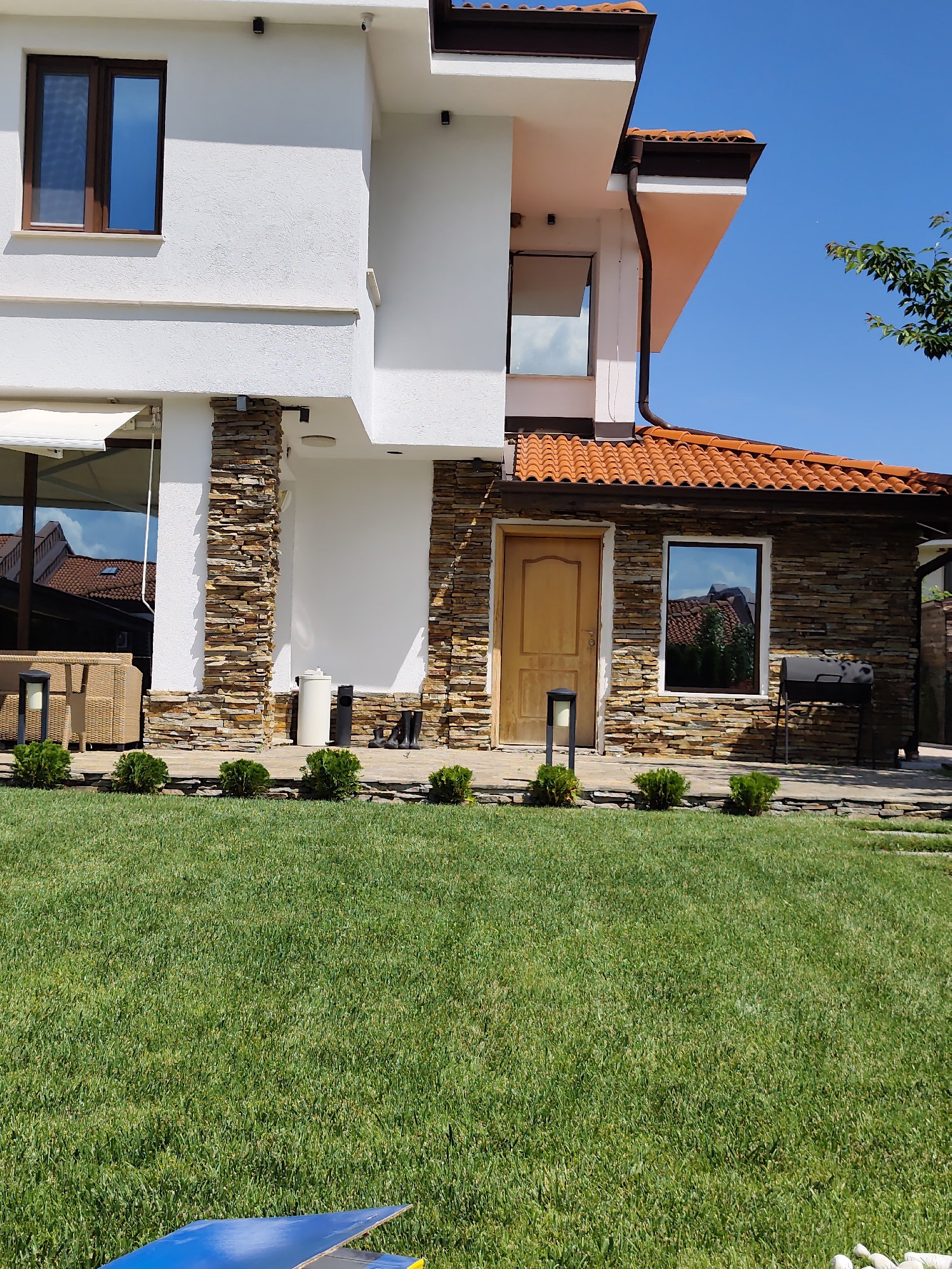 The image shows a modern house with a stone facade and a terracotta tiled roof. The house is surrounded by a well-maintained green lawn and a stone pathway. In front of the house, there are several small, manicured bushes. On the patio area, there is outdoor furniture, a grill, a wooden door, and several pairs of black boots. The sky is clear and blue. The location is specified as Kladnitsa, Bulgaria.