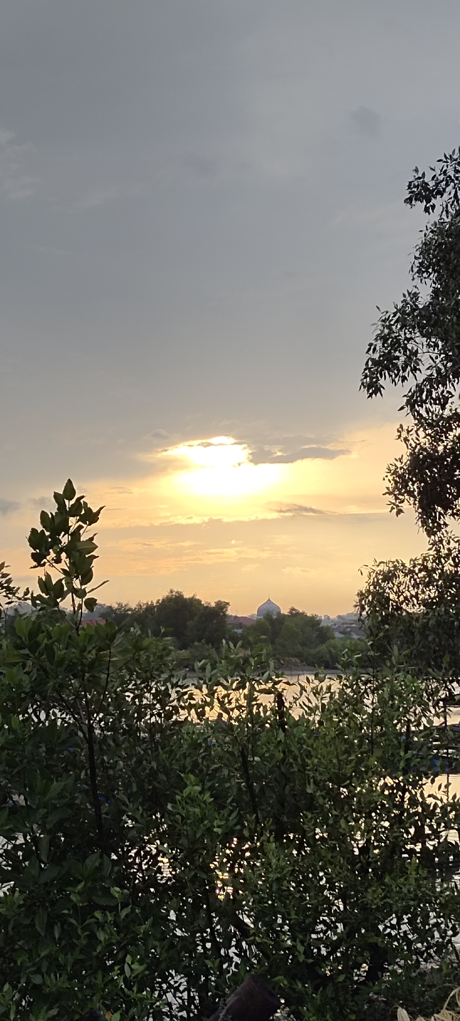 The image depicts a serene sunset scene over a body of water, likely in Lhokseumawe, Indonesia, given the presence of a domed building resembling a mosque in the distance. The foreground is dominated by lush green foliage, possibly mangrove trees, which partially obscure the view of the water and the settlement beyond. The sky is a blend of soft grays and warm yellows, with the sun partially hidden behind clouds, casting a gentle glow on the water's surface. The overall impression is one of tranquility and natural beauty.