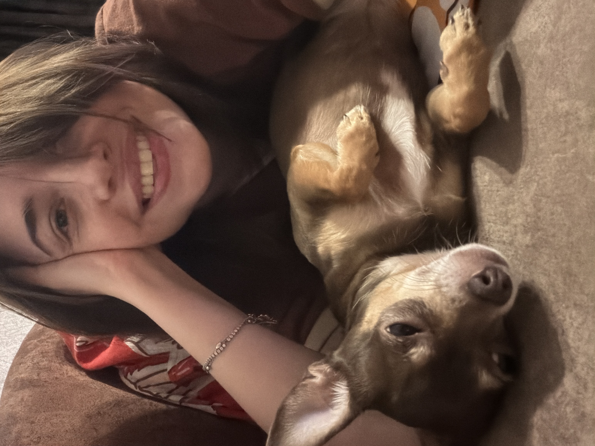 A person with short, brown hair, lying on their side, with their right arm propped up on their head, supports their head with their hand. The person is smiling, with their mouth open, showing their teeth. A small, brown dog is lying on its back in front of the person, with its paws in the air. The dog's eyes are partially closed, and it appears to be relaxed. The background is blurred, but it appears to be a couch or bed with a patterned blanket. The scene is dimly lit, with soft lighting.