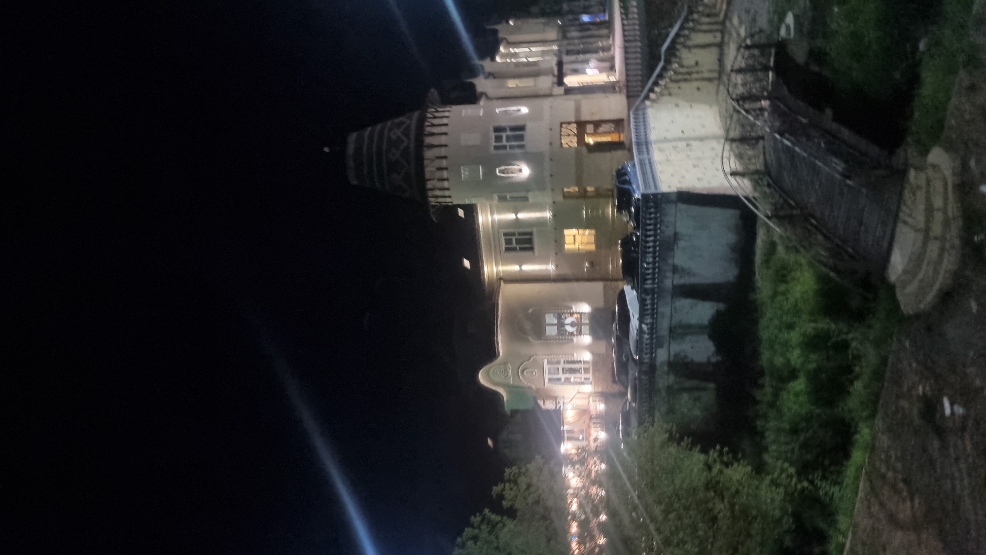 The image depicts a building with a tower at night in Berehove, Ukraine. The building is illuminated with exterior lights, highlighting its architectural features, including multiple windows and a decorative top on the tower. In front of the building, there is a row of parked cars. A wooden footbridge arches over a grassy area in the foreground, leading to a textured wall. Trees and foliage are visible on the left side of the image. The overall scene suggests an outdoor setting at an event or establishment, possibly a restaurant or hotel, given the lighting and the presence of parked vehicles.