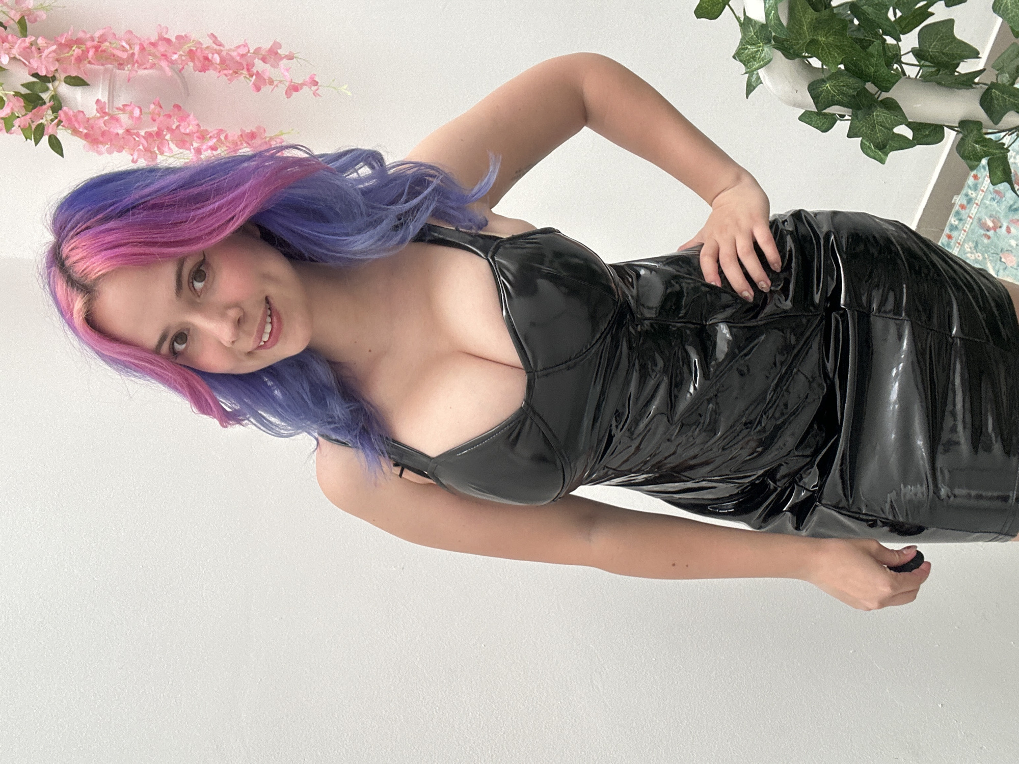 A young woman with vibrant, dual-toned pink and purple hair is captured in a full-body portrait, set against a bright, minimalist background. Her long, wavy hair, transitioning from pink at the roots to purple at the ends, frames her face as she smiles warmly, looking directly at the viewer. She is wearing a form-fitting, black mini-dress with a high-shine finish, possibly made of latex or PVC, featuring a sweetheart neckline and thick shoulder straps.
The woman is posing with her right hand resting on her hip, while her left hand is down by her side, holding a small, dark, spherical object. Her posture suggests confidence and playfulness. The background consists primarily of a plain white wall, which provides a clean contrast to her colorful hair and dark dress. Decorative elements are visible in the upper corners: cascading pink artificial flowers are on the upper left, and green artificial ivy or similar foliage is seen on the upper right, suggesting an indoor setting with decorative plants. A small section of a patterned rug, featuring blue and white tones, is visible at the very bottom right of the frame. The lighting is bright and even, indicating either a well-lit indoor environment or a daytime setting. There is no visible text in the image. The photo was taken in the City of Zagreb, Croatia.