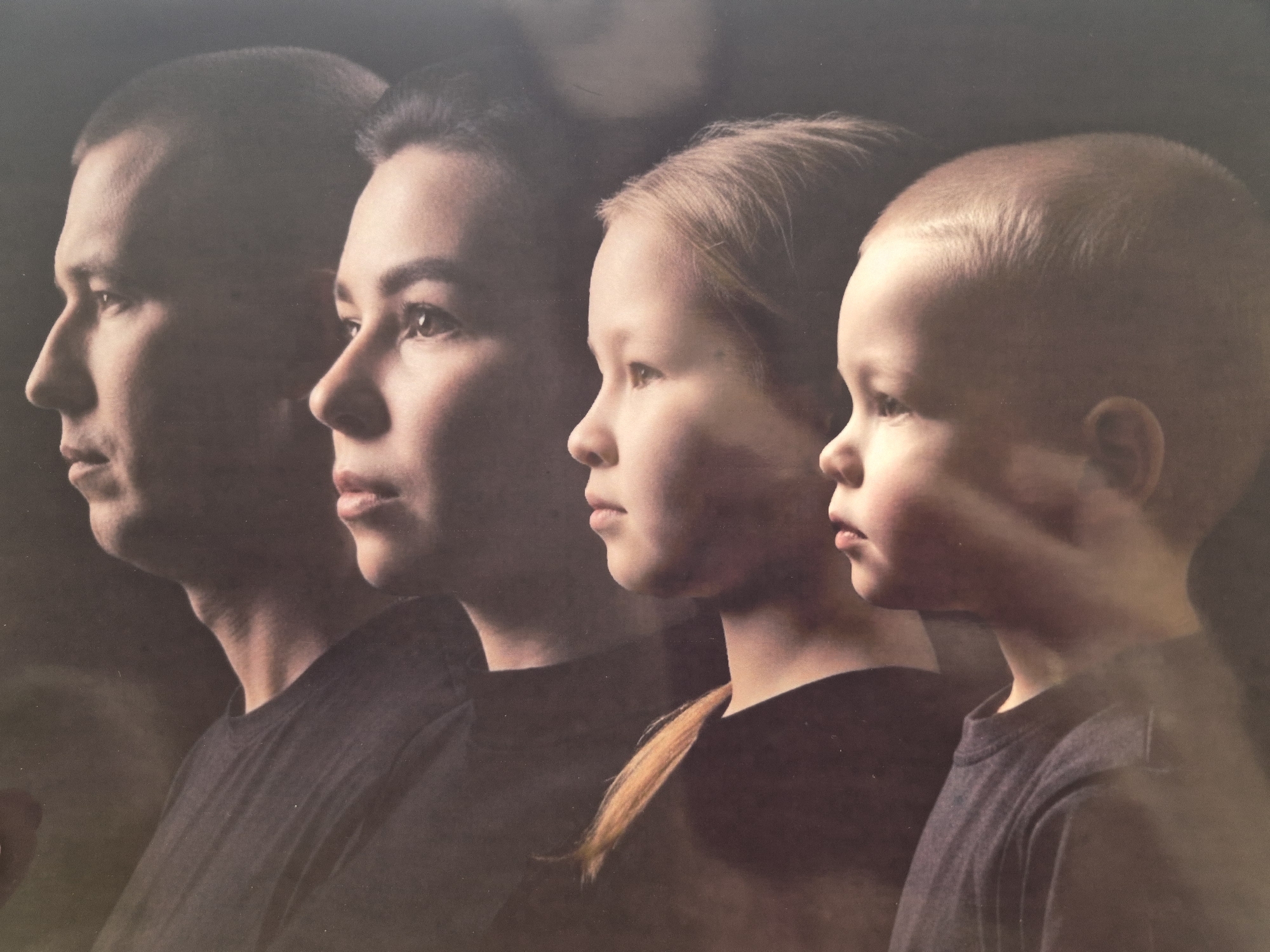 This image is a profile portrait featuring four individuals, arranged from left to right, appearing to represent a family across generations. The subjects are positioned looking towards the left, with the background uniformly dark and out of focus.
On the far left is an adult male with short, dark hair and a serious or contemplative expression. He wears a dark, possibly black, t-shirt. To his right and slightly overlapping is an adult female, also with dark hair pulled back, and a similar neutral expression. She wears a dark t-shirt.
Next is a young girl with blonde hair, which is styled in a ponytail that extends down her back. Her profile is clear, and she also maintains a calm, neutral gaze to the left. She is wearing a dark t-shirt.
On the far right is a very young child, likely a toddler or young boy, with light brown or blonde hair. His profile shows soft facial features, and he too looks to the left. He is dressed in a dark grey or black t-shirt.
The lighting strongly illuminates the subjects' profiles, creating a sense of depth and highlighting their features against the dark, indistinct background. No specific objects, text, or location cues are visible within the frame, beyond the implied indoor setting suggested by the studio-like lighting and background. The overall mood is serene and reflective.